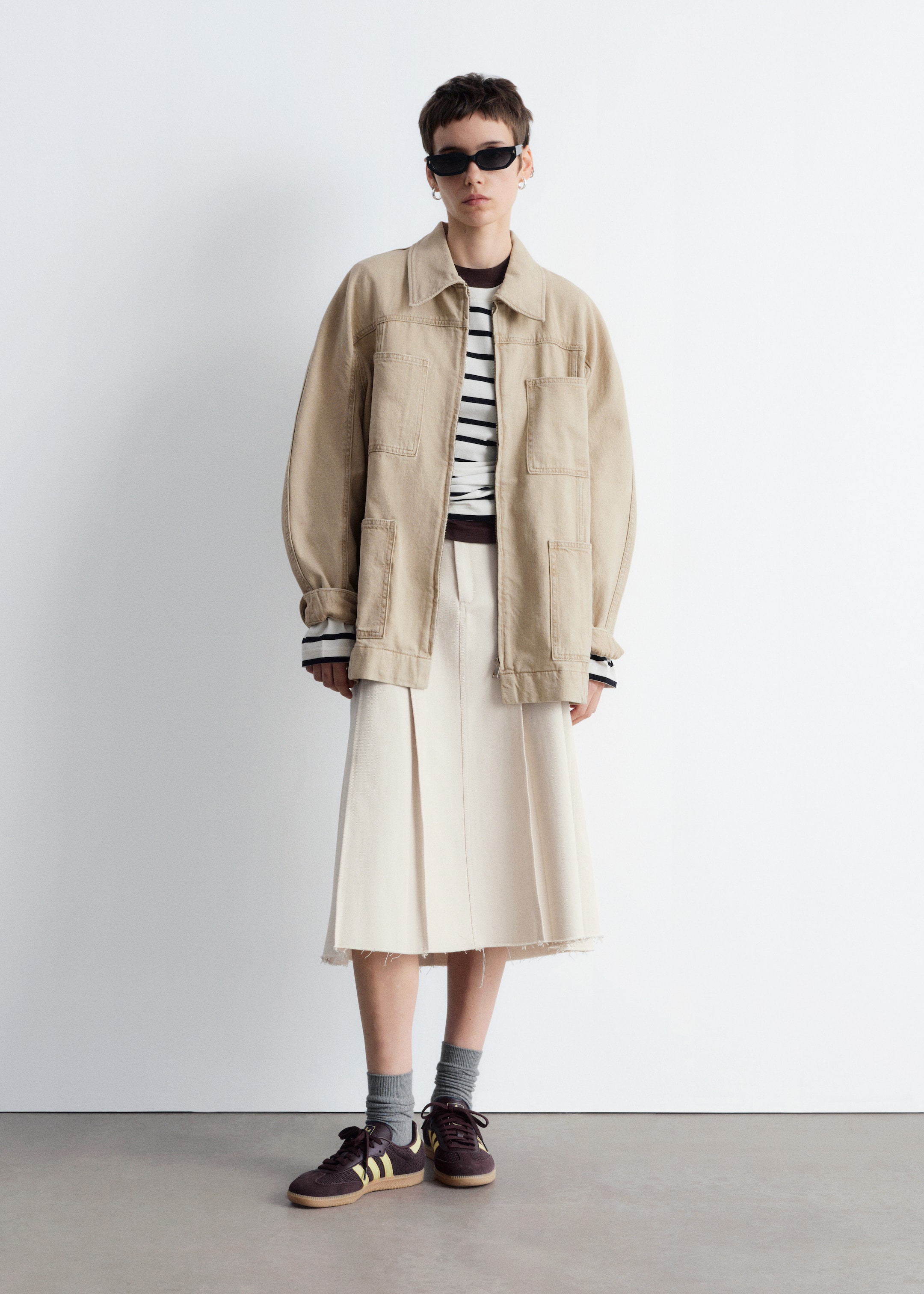 Agrandir l'image: Une veste de travail beige surdimensionnée, avec un col pointu et plusieurs poches à rabat, est portée ouverte sur un haut à manches longues rayé noir et blanc superposé d'un vêtement marron. Une jupe midi plissée de couleur crème avec un ourlet brut est portée en dessous, assortie à des baskets marron ornées de rayures jaunes et de chaussettes grises visibles.