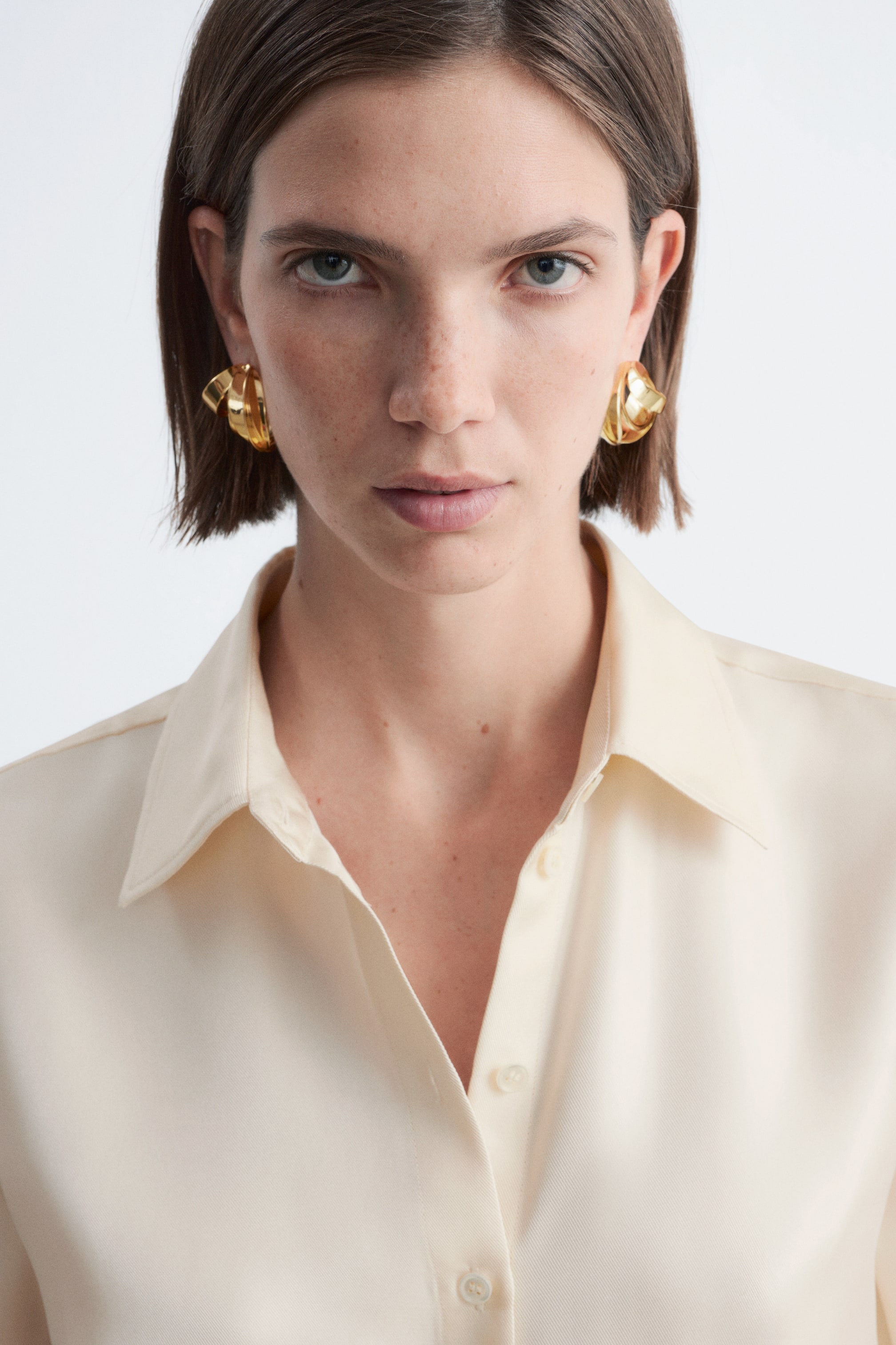 View larger image: A pair of gold-toned earrings is being worn, featuring a sculptural, folded design. Each earring sits close to the earlobe, showcasing its abstract, curved surfaces.