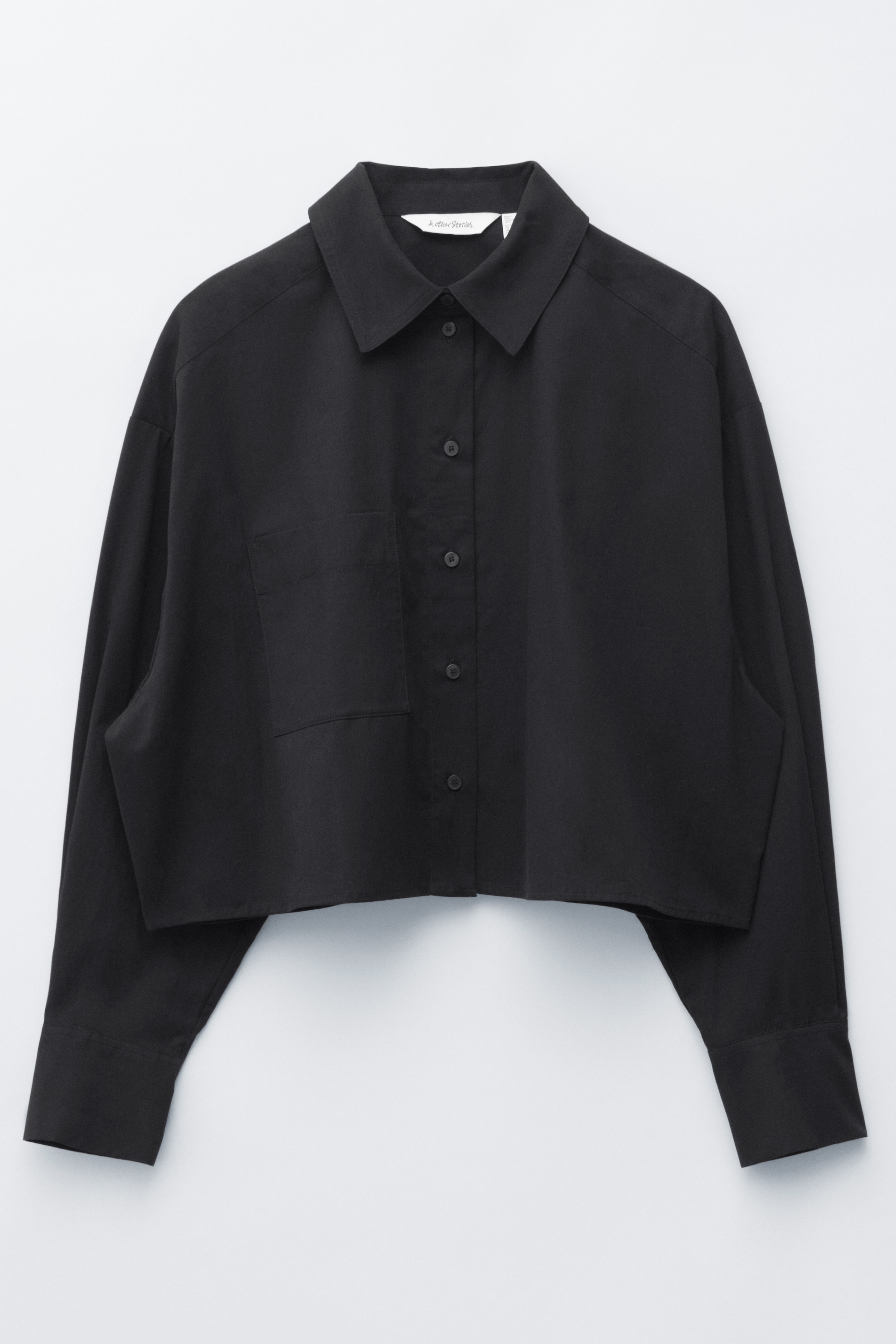 Ver imagen más grande: Se presenta una camisa negra de corte amplio y recortado vista desde el frente. Cuenta con un clásico cuello vuelto, una etiqueta de marca visible en el interior del escote, una tapeta abotonada con botones oscuros, un bolsillo de parche único en el pecho izquierdo y mangas largas con puños abotonados.