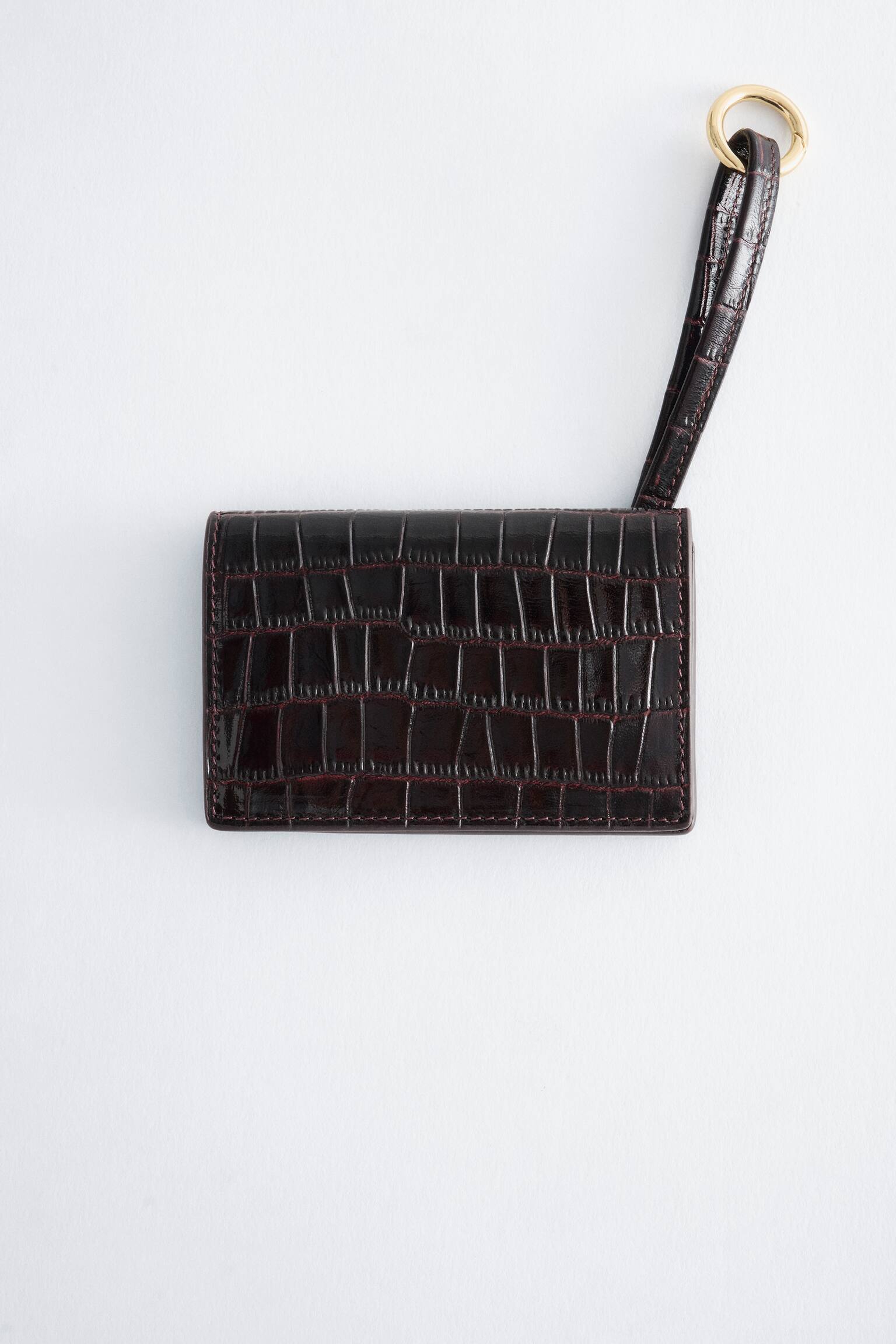 A dark burgundy, glossy croc-effect leather cardholder is facing front, showcasing its textured rectangular body with tonal stitching. A matching croc-effect wrist strap extends upward from the top-right corner, ending in a circular, gold-toned metal ring.