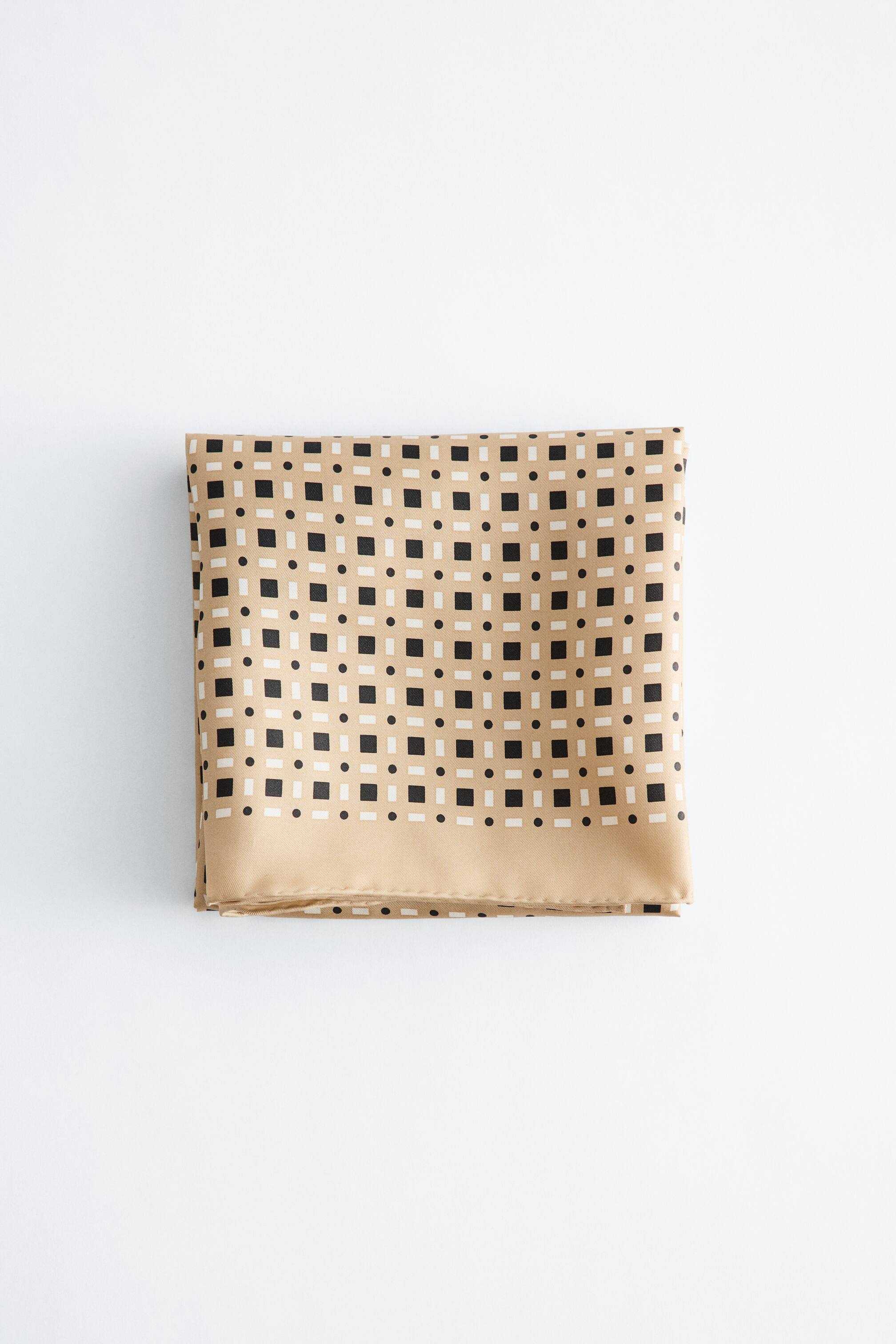 הצגת תמונה גדולה יותר: צעיף מקופל לצורת מלבן, מציג בסיס בצבע בז' עם דפוס גיאומטרי חוזר של ריבועים שחורים קטנים וקווים מלבניים לבנים קטנים. גבול בצבע בז' אחיד נראה לאורך הקצה התחתון.