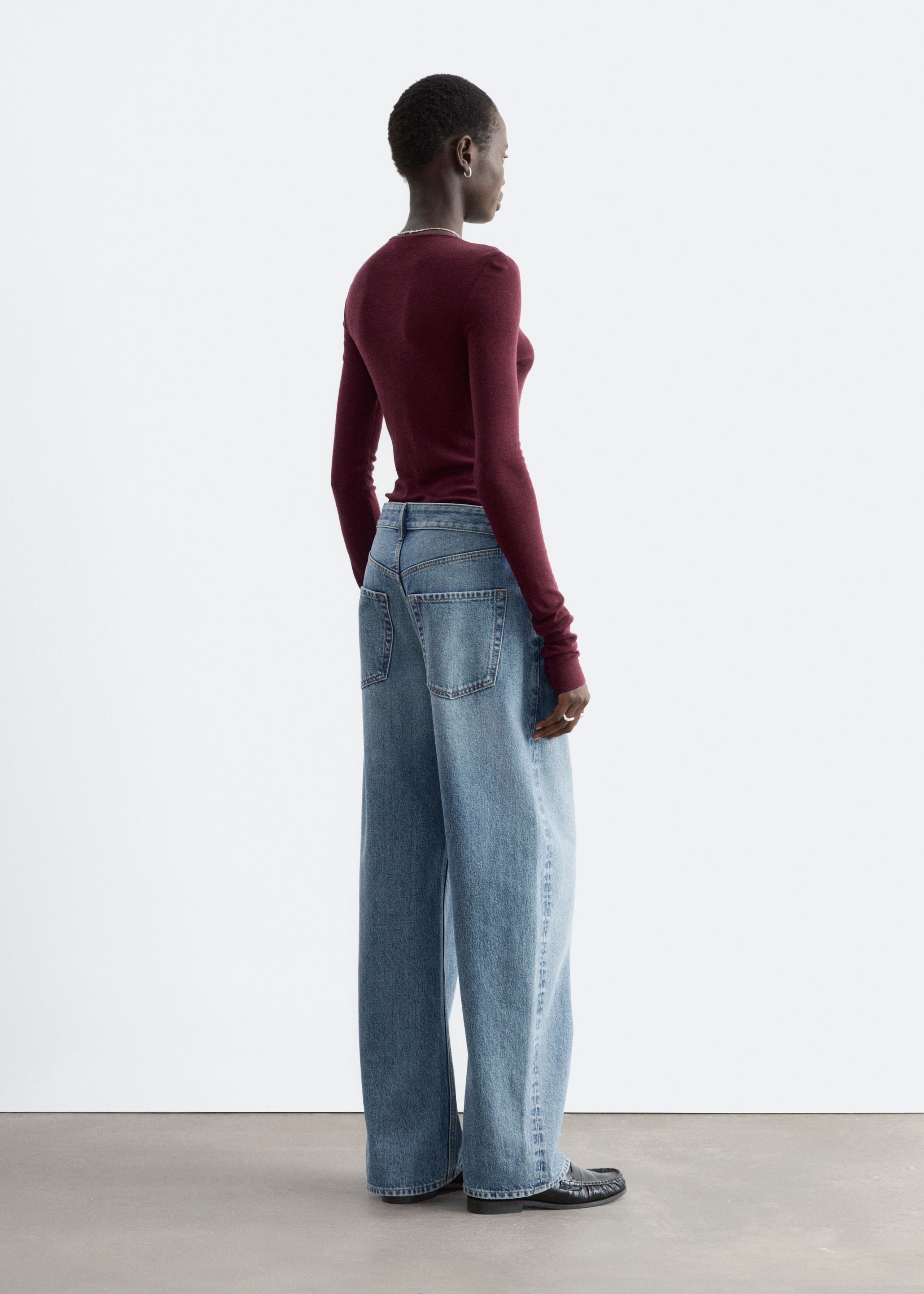 Ein bordeauxrotes, glatt gestricktes Oberteil aus Wolle wird vom Model getragen und zeichnet sich durch eine schmale Passform und lange Ärmel aus. Das Oberteil ist in eine blaue Wide-Leg-Jeans gesteckt und hebt aus einer Rückenansicht in dreiviertel Perspektive seine feine Strickstruktur hervor.