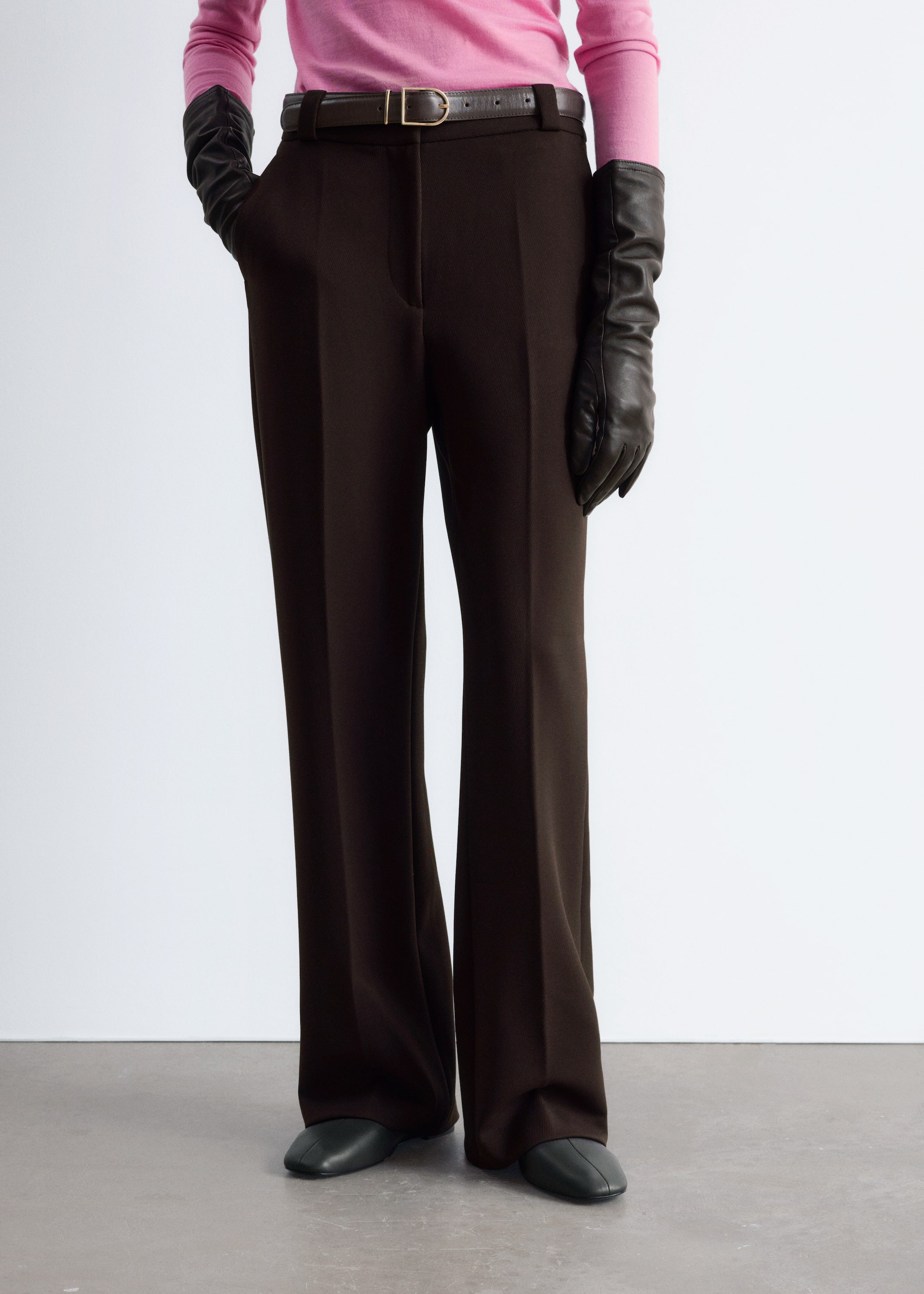 Agrandir l'image: Un pantalon large marron foncé est porté par le mannequin, avec des plis marqués distincts et des poches avant en biais. Une ceinture marron foncé est portée à travers les passants de ceinture, et l'une des mains gantées du mannequin est placée dans une poche avant.