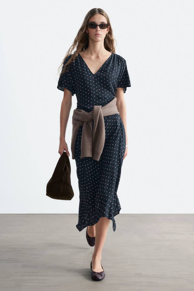 Ein dunkelblaues Midikleid mit rosa Punkten wird von dem Model getragen und verfÃ¼gt Ã¼ber einen V-Ausschnitt, kurze Ãrmel und dezente Raffungen an den Schultern und der Taille. Um die Taille des Kleides ist ein brauner Pullover gebunden, und das Model trÃ¤gt eine dunkelbraune Handtasche. - 1
