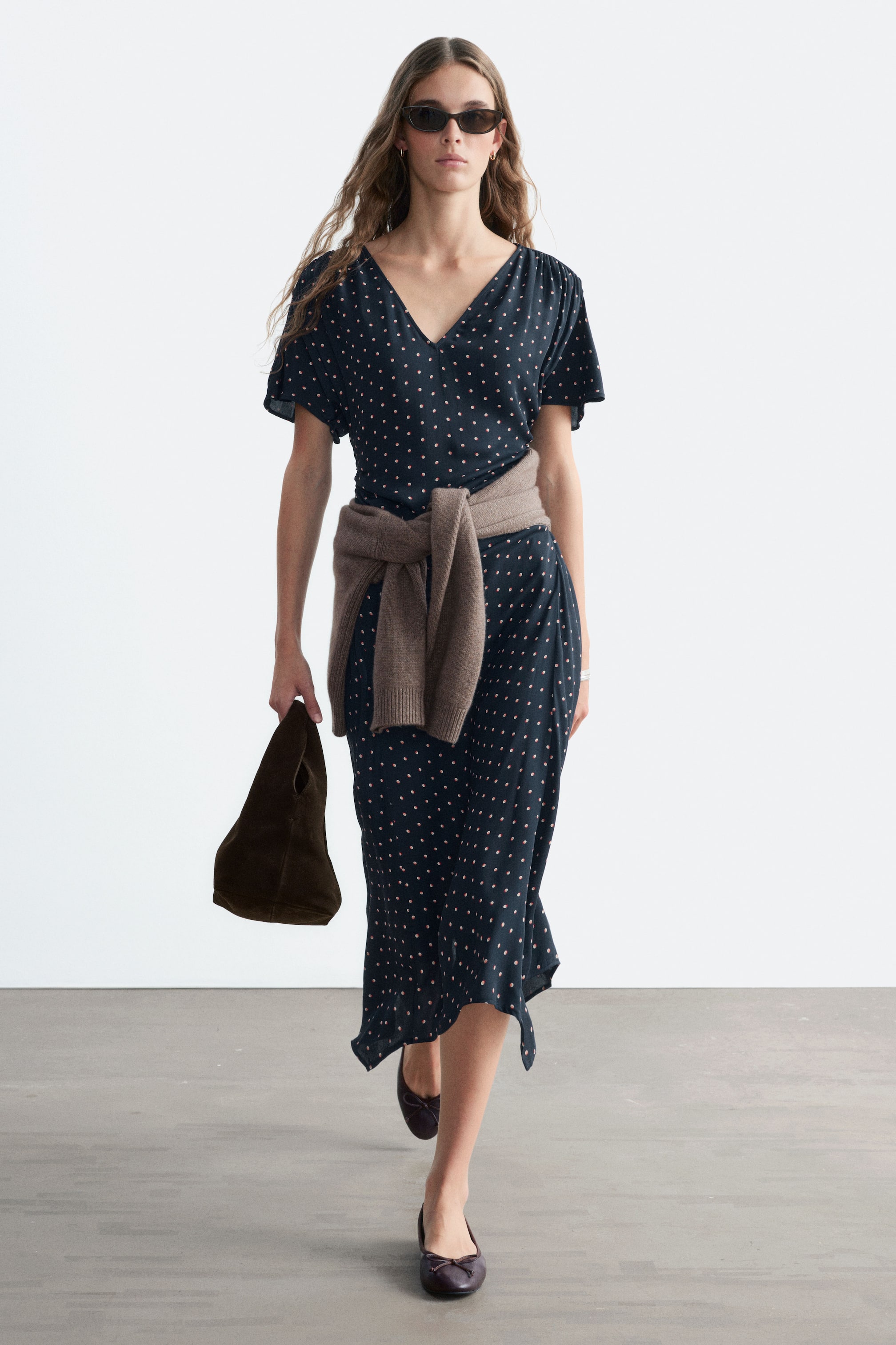 Agrandir l'image: Une robe midi bleu foncé à pois roses est portée par le mannequin, avec un col en V, des manches courtes et de légers fronces aux épaules et à la taille. Un pull marron est noué à la taille de la robe, et le mannequin porte un sac à main marron foncé.