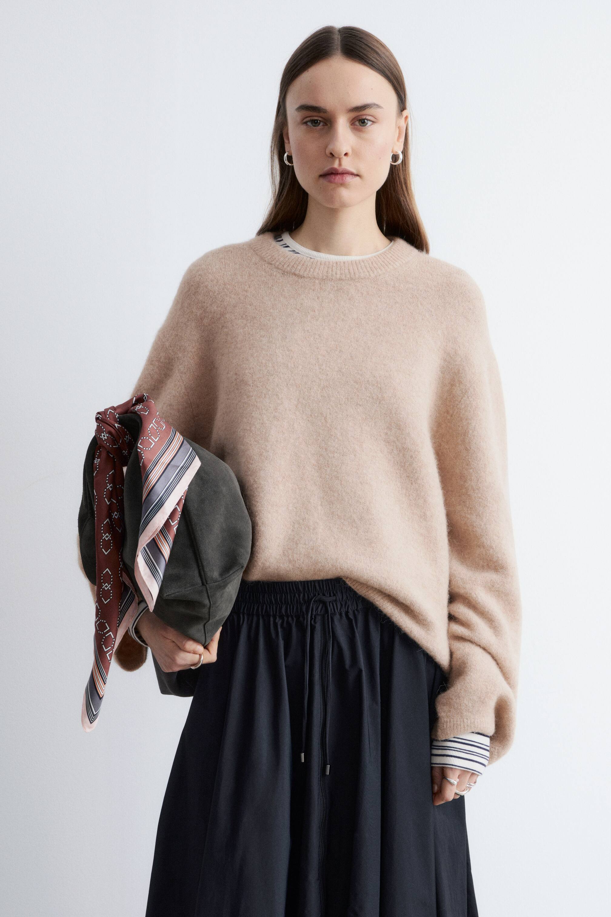 Преглед на по-голямо изображение: Бежов пухкав плетен пуловер с кръгло деколте, който се отличава с релаксирана кройка и дълги ръкави, е показан отпред, с видим бял слой на деколтето и раирани маншети, които се простират от ръкавите му. Той е съчетан с черна миди пола, която се отличава с еластична талия с връзка.