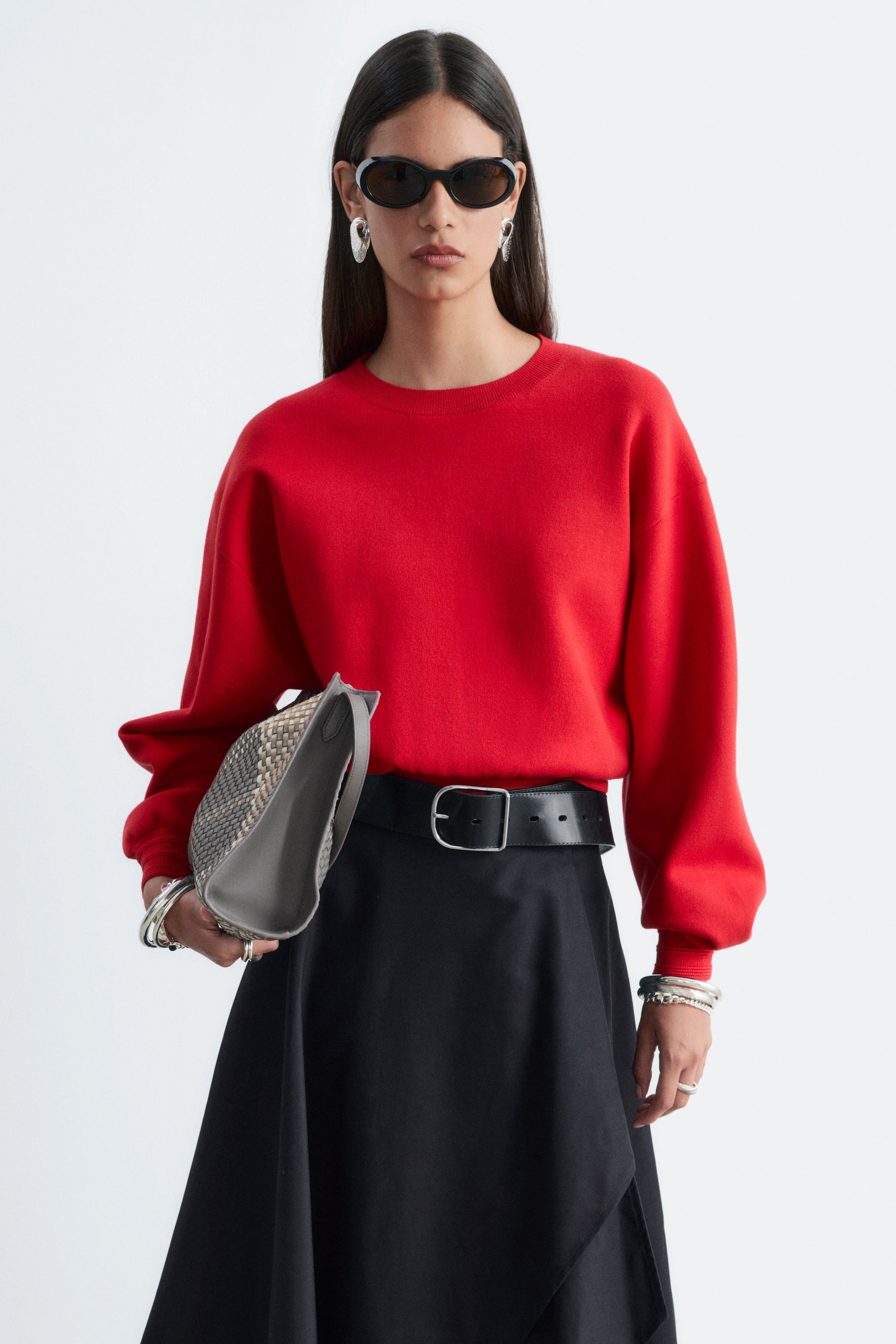 Größeres Bild anzeigen: Ein roter gestrickter Pullover mit Rippbündchen wird vom Model getragen und verfügt über einen Rundhalsausschnitt sowie locker geschnittene Ärmel mit Rippbündchen. Der Pullover ist in einen schwarzen Rock mit einem breiten schwarzen Gürtel gesteckt.