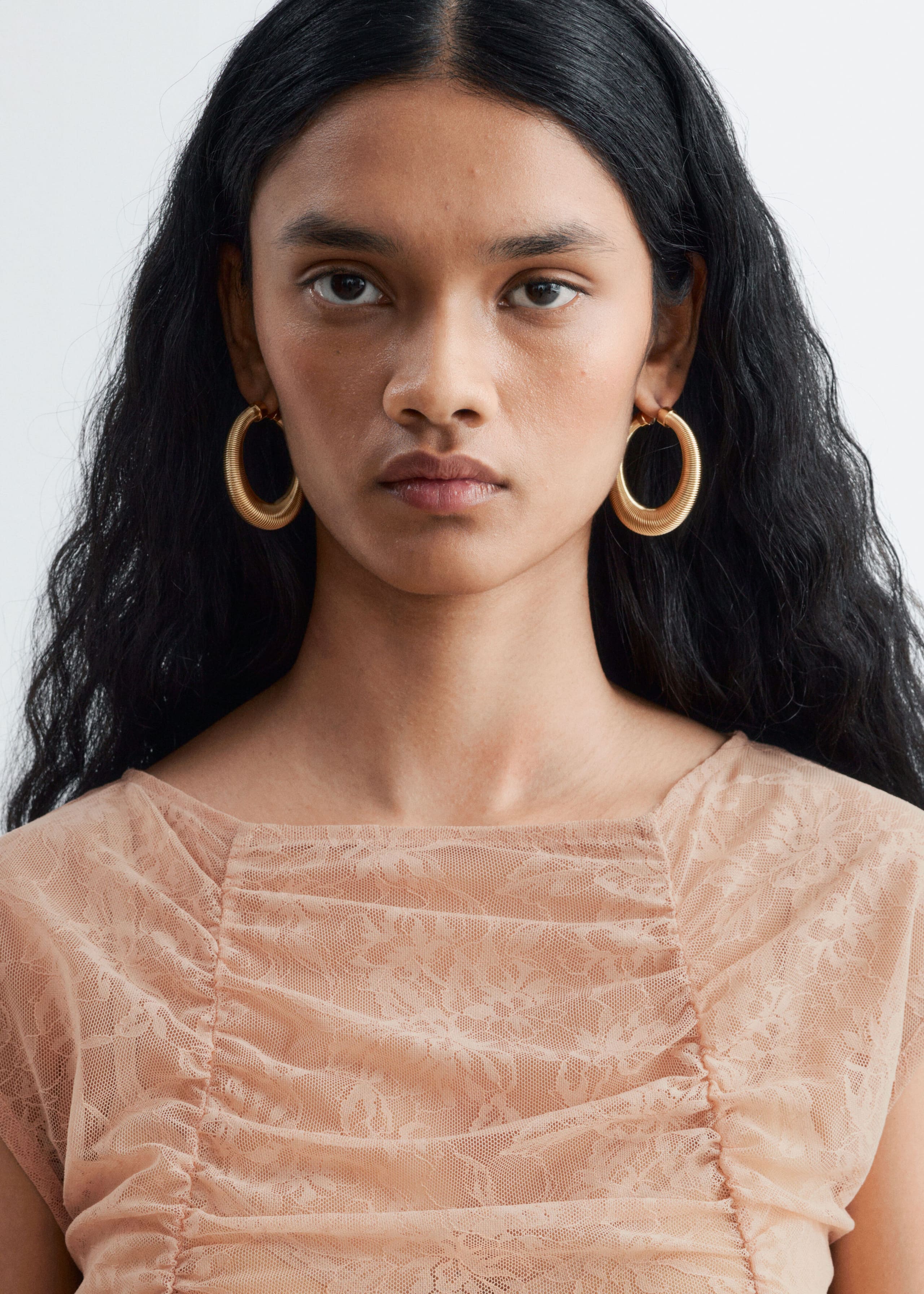 A light peach lace top featuring a boat neckline and ruched detailing is being worn by the model. The close-up shows the gathered floral lace fabric across the chest, styled with large gold hoop earrings.
