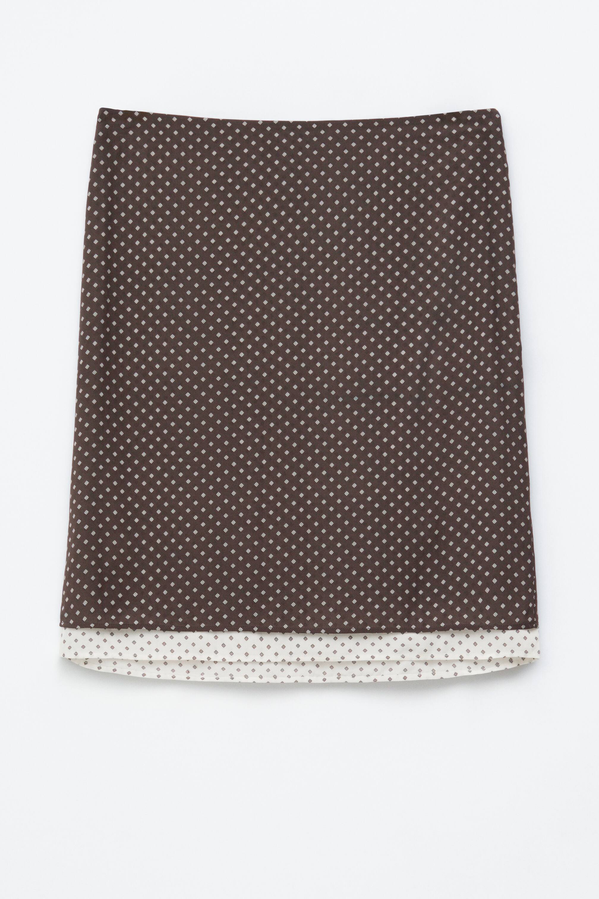 Преглед на по-голямо изображение: Тъмнокафява пола с предно лице, която се отличава с повтарящ се модел от малки, светли ромбовидни форми. Полата също така има контрастиращ кремав кант в долния подгъв, който е украсен с малки ромбове.