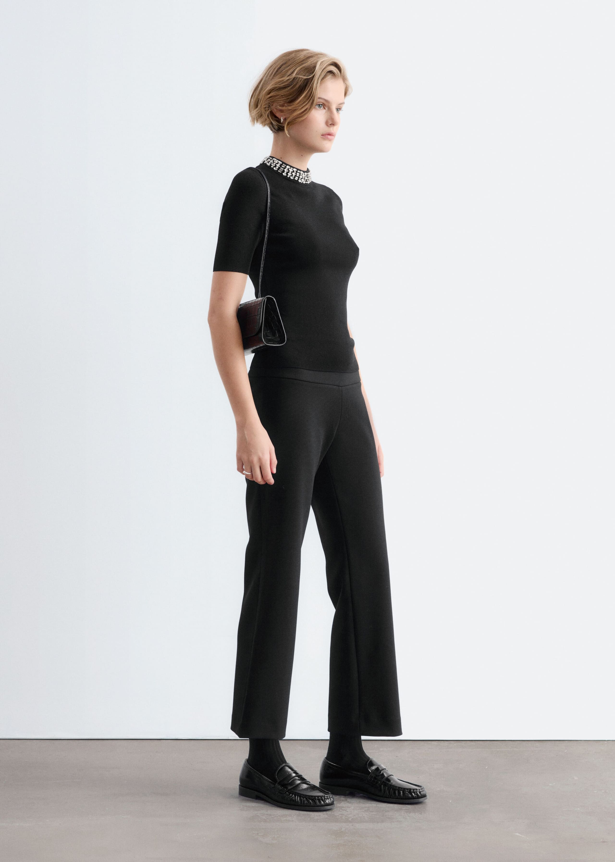 The model is wearing a black short-sleeve fitted top with a mock neck embellished with rows of silver rhinestones. The top is styled with coordinating black trousers and a small black shoulder bag.