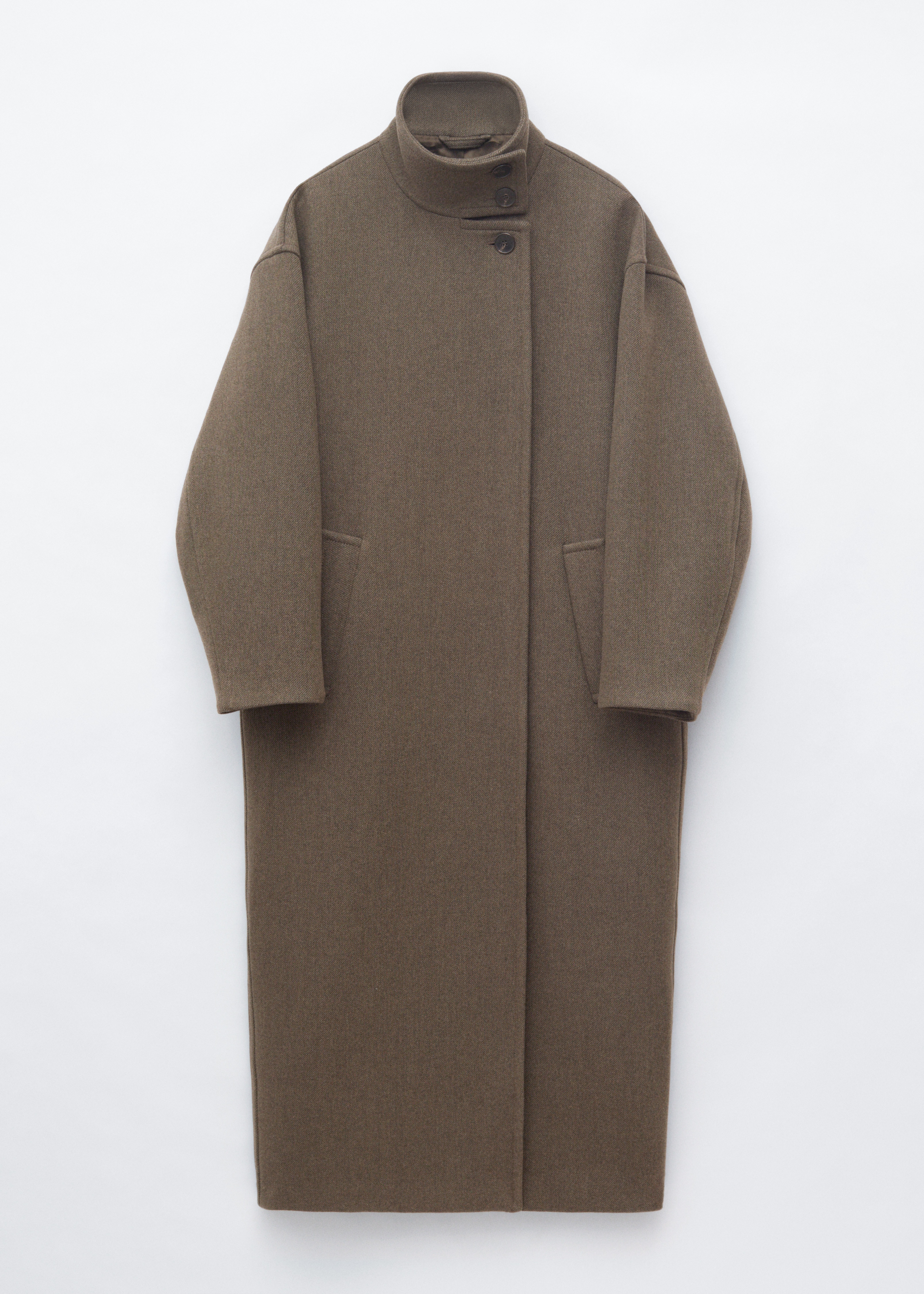 Een oversized wollen jas tot op de enkels in een donker olijfbruin kleur wordt gedragen door het model, met brede revers en steekzakken. De jas is gelaagd over een donkerbruine cardigan met knopen en een top met hoge hals, waarbij het model Ã©Ã©n hand in een jaszak houdt en een donkere totebag draagt.