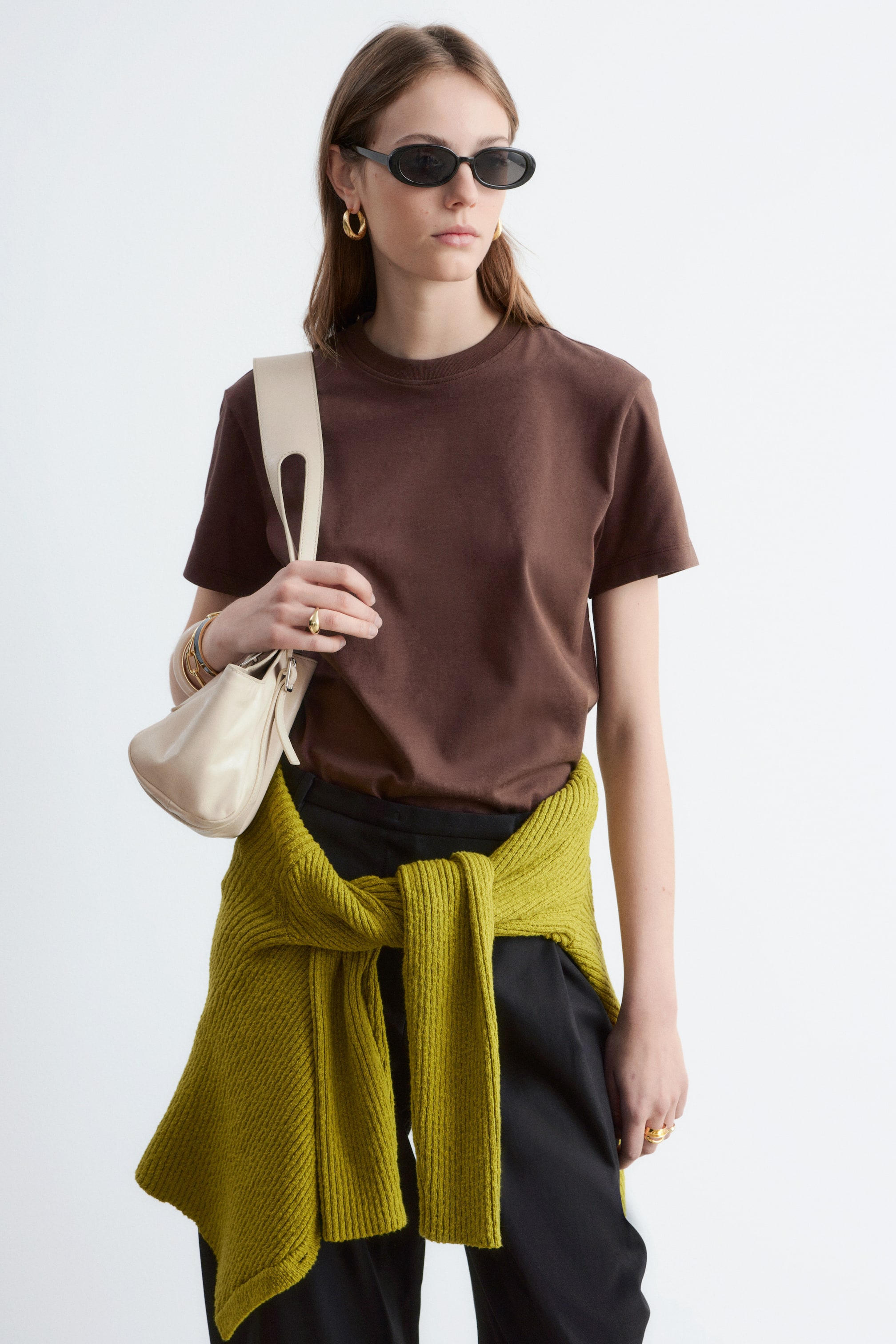 Grösseres Bild ansehen: Ein braunes T-Shirt mit Rundhalsausschnitt und kurzen Ärmeln wird vom Model getragen. Das T-Shirt ist in eine schwarze Hose gesteckt, und ein limettengrüner Pullover ist um die Taille des Models gebunden.