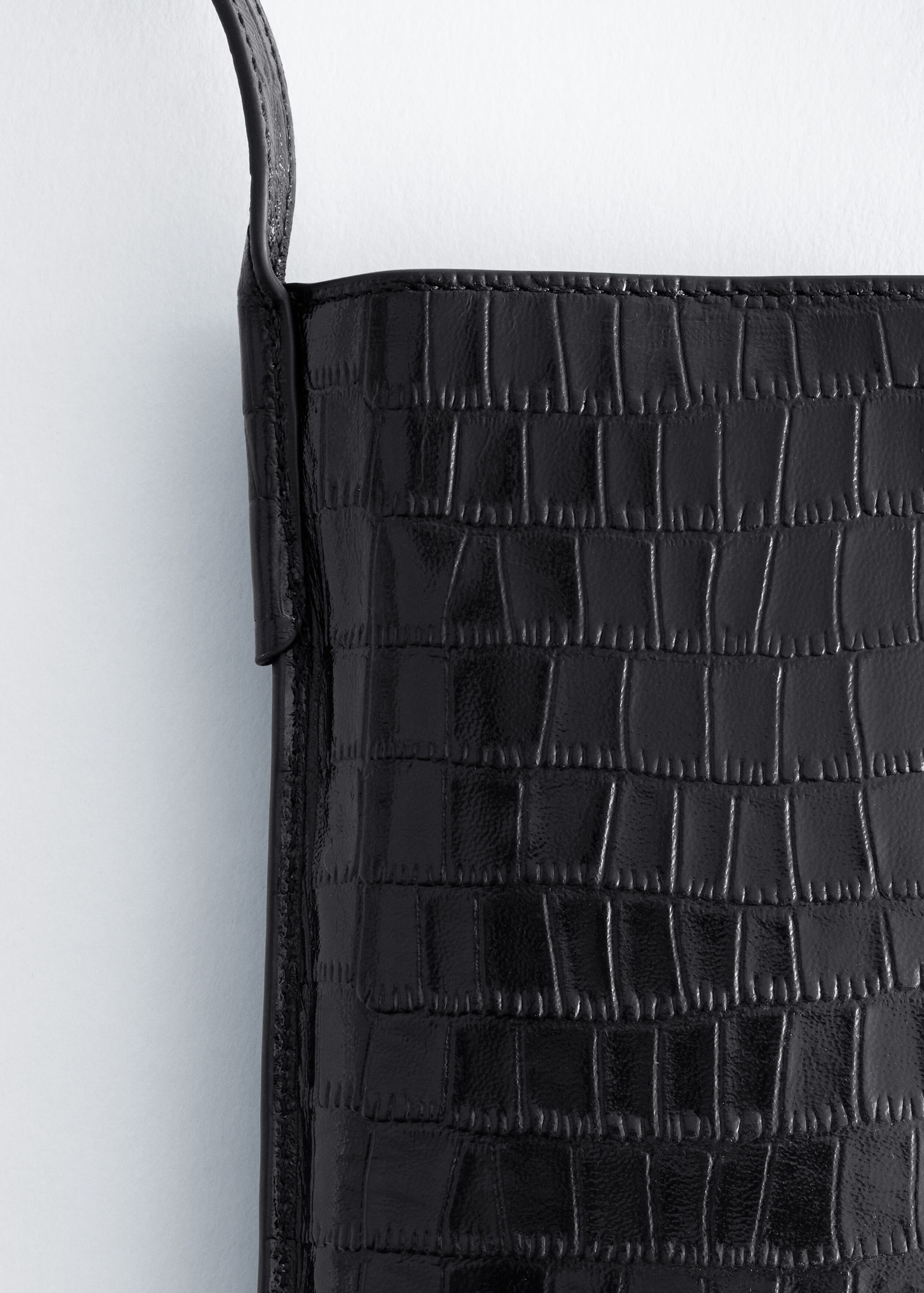 Un sac pour téléphone noir à relief façon crocodile est présenté en gros plan, mettant en valeur sa finition brillante et texturée ainsi que ses coutures nettes le long du bord supérieur. Une fine bandoulière assortie est fixée sur le côté supérieur gauche, soulignant la structure minimaliste du sac.