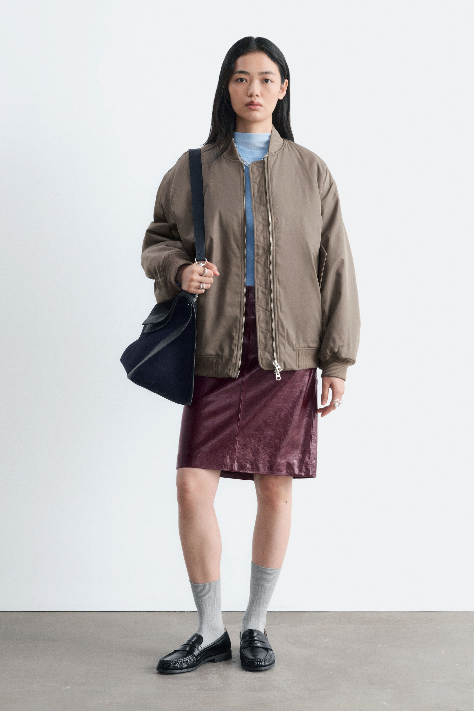 Agrandir l'image: Une longue veste bomber dans une teinte taupe brun est portée ouverte par le mannequin, mettant en valeur un col côtelé, des poignets côtelés et une fermeture zippée sur le devant. La veste est superposée à un top bleu clair à col montant et à une jupe en cuir bordeaux, tandis qu’un sac porté épaule noir est porté sur l’épaule du mannequin.