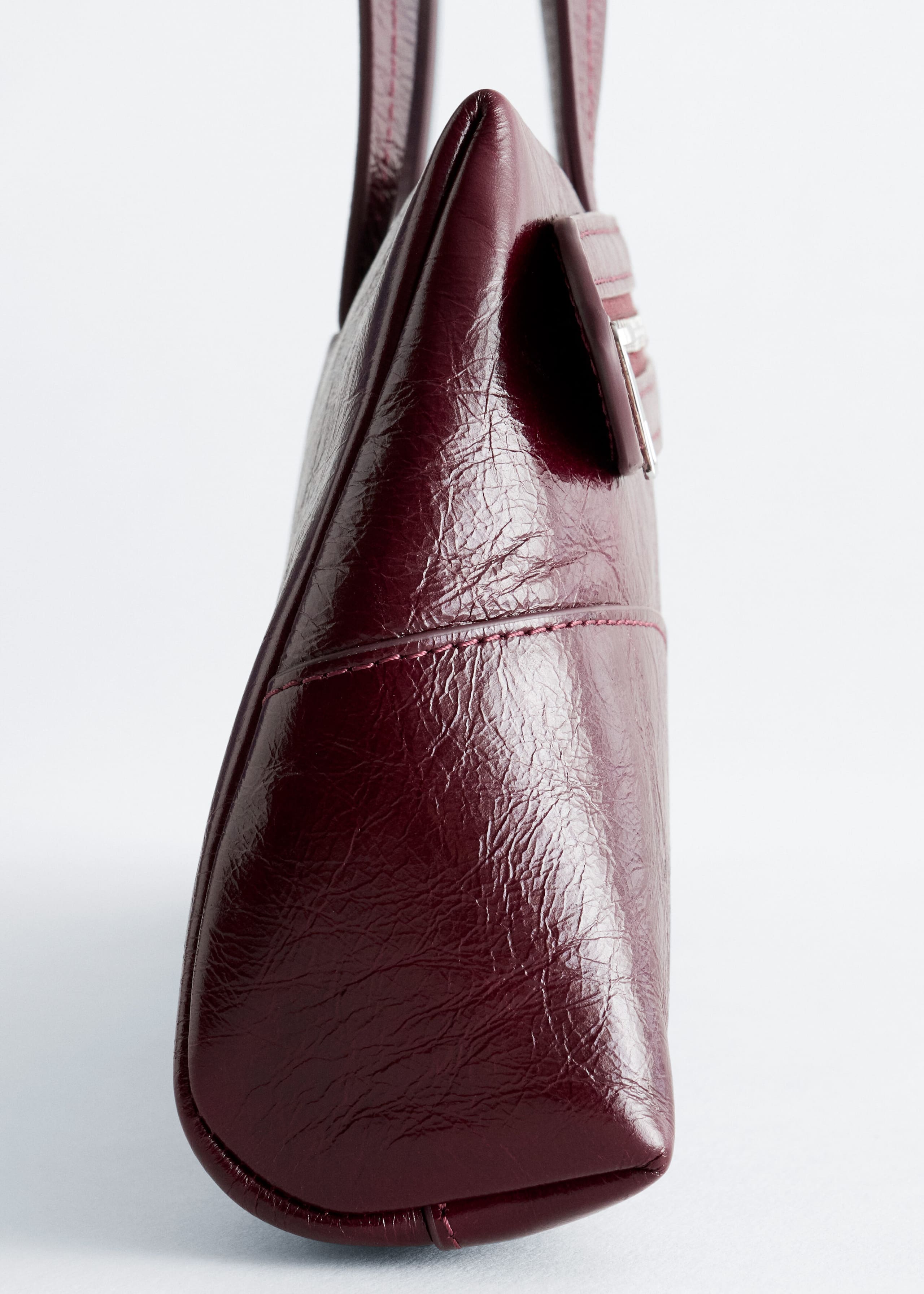 A burgundy shoulder bag is shown in side profile, highlighting its glossy, crinkled leather finish and tonal stitching details. Two flat shoulder straps extend upwards from the top edge of the bag’s tapered silhouette.