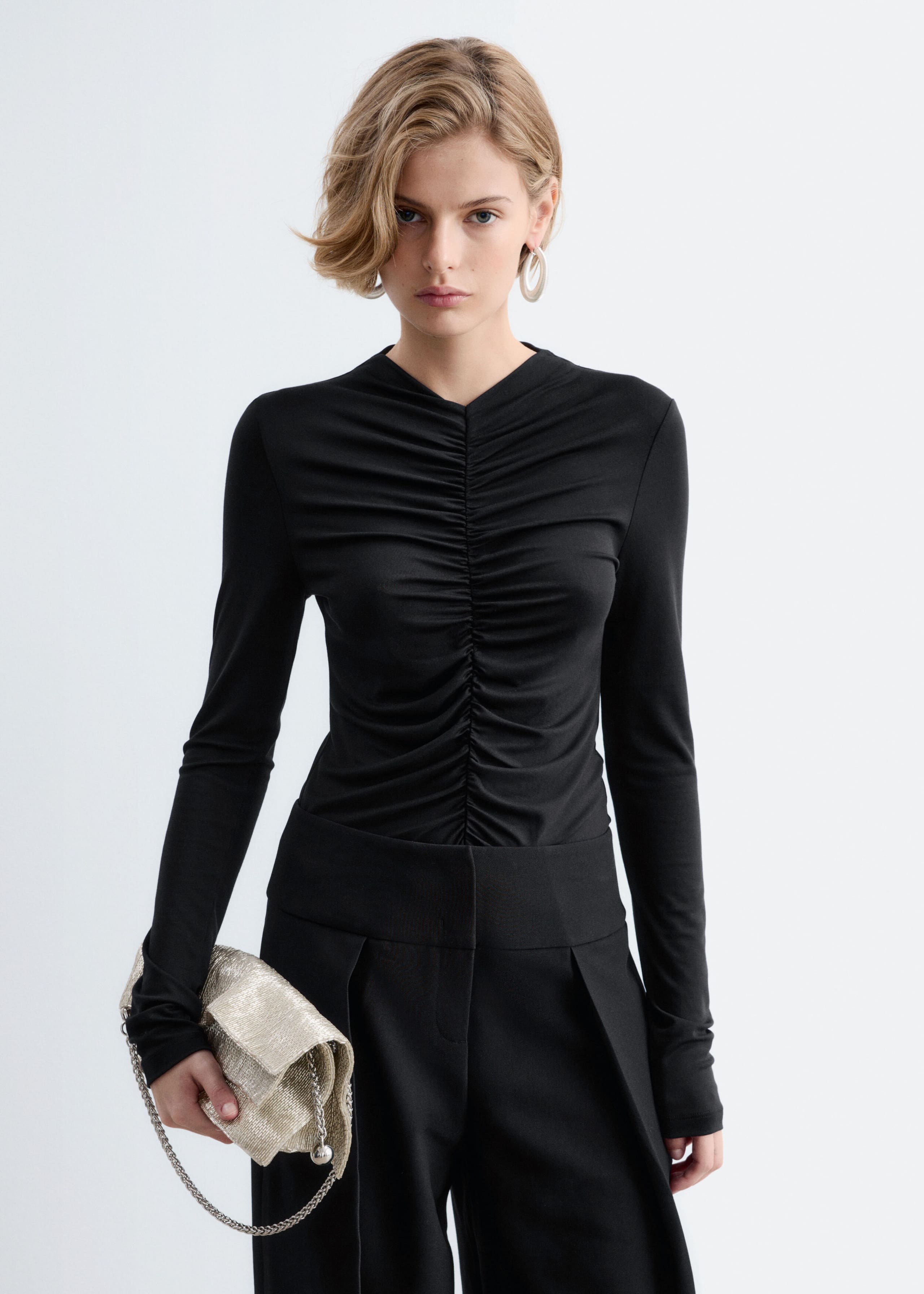 A black long-sleeve top featuring prominent gathers along the center-front seam is being worn by the model. The fitted top is styled with black trousers, and the model is holding a textured gold clutch bag.