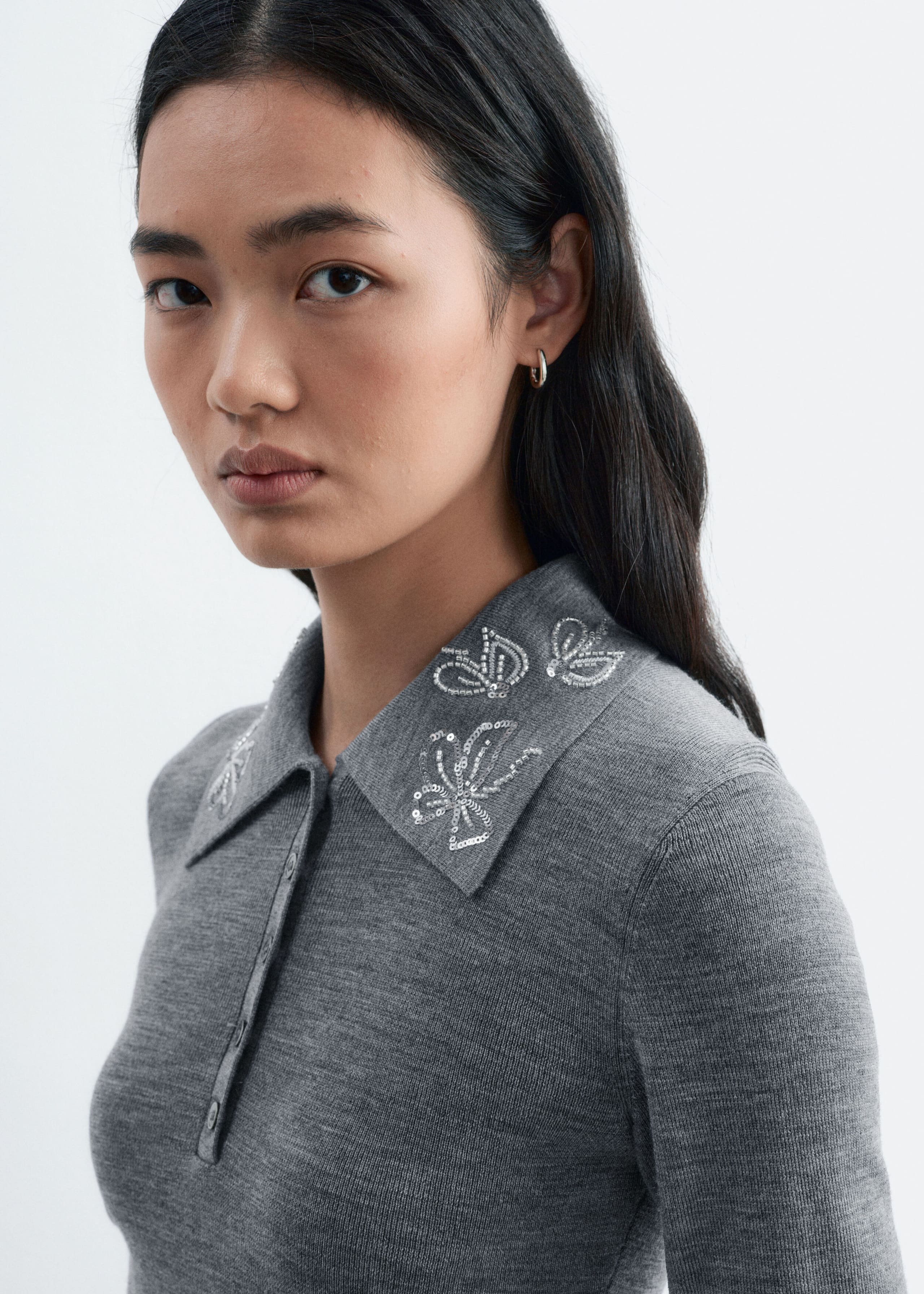 Ein grauer Woll-Polo-Pullover mit geknöpfter Frontblende wird vom Model getragen. Der spitze Kragen ist mit Schmetterlingsstickerei mit silbernen Pailletten verziert.
