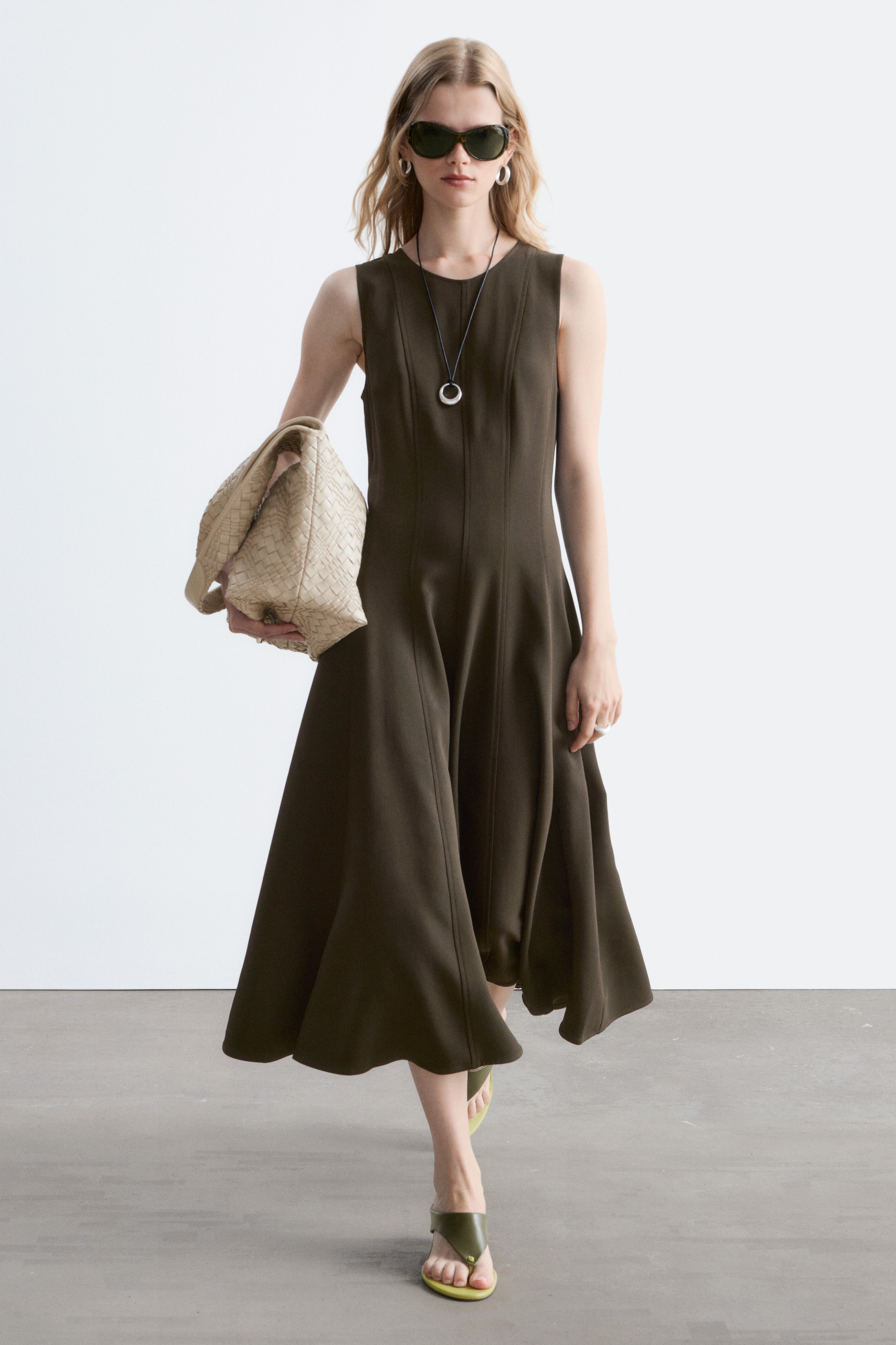 Agrandir l'image: Une robe midi sans manches vert olive présente un corsage ajusté avec des coutures verticales qui se prolongent en une jupe évasée et fluide. Un long collier noir avec un pendentif circulaire argenté est porté par-dessus la robe, et un sac léger en tissu tressé est porté à côté.