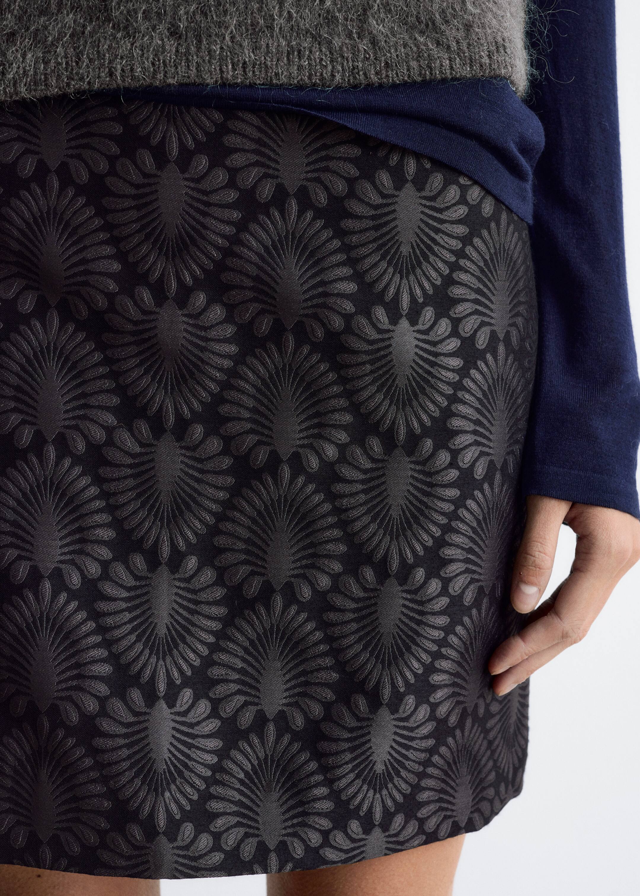 Größeres Bild ansehen: Ein schwarzer Rock wird aus einem leichten Winkel gezeigt, mit einem sich wiederholenden, Ton-in-Ton geprägten Muster von stilisierten, fächerartigen Motiven. Der Rock wird in der Taille getragen, mit einem dunkelblauen langärmeligen Oberteil und einem grauen Pullover darüber.