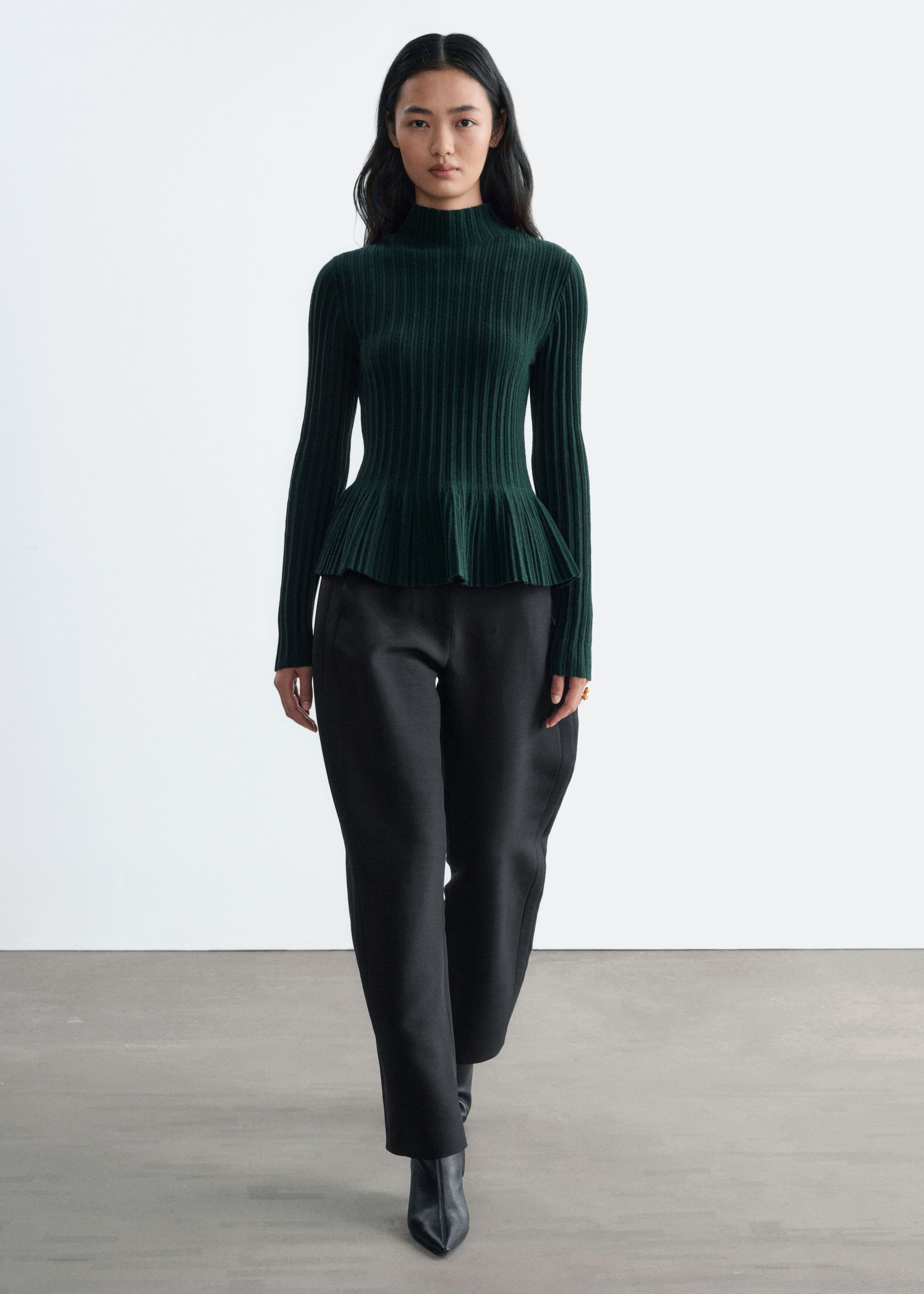 A dark green, ribbed-knit top featuring a mock neck, long sleeves, and a flared peplum hem is being worn by the model. The top is styled with black trousers and black pointed boots.