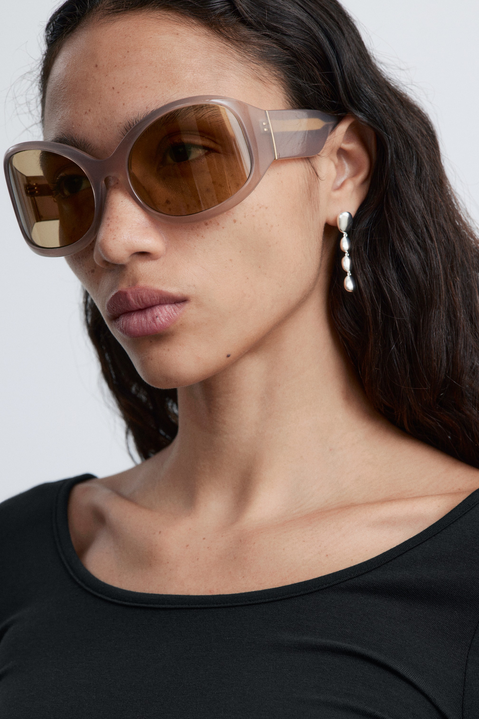 Ver imagem maior: Os óculos de sol estão a ser usados, ligeiramente inclinados para o lado, apresentando armações retangulares arredondadas e de grandes dimensões, num tom translúcido de taupe, equipados com lentes castanhas e exibindo um discreto detalhe dourado nas hastes.