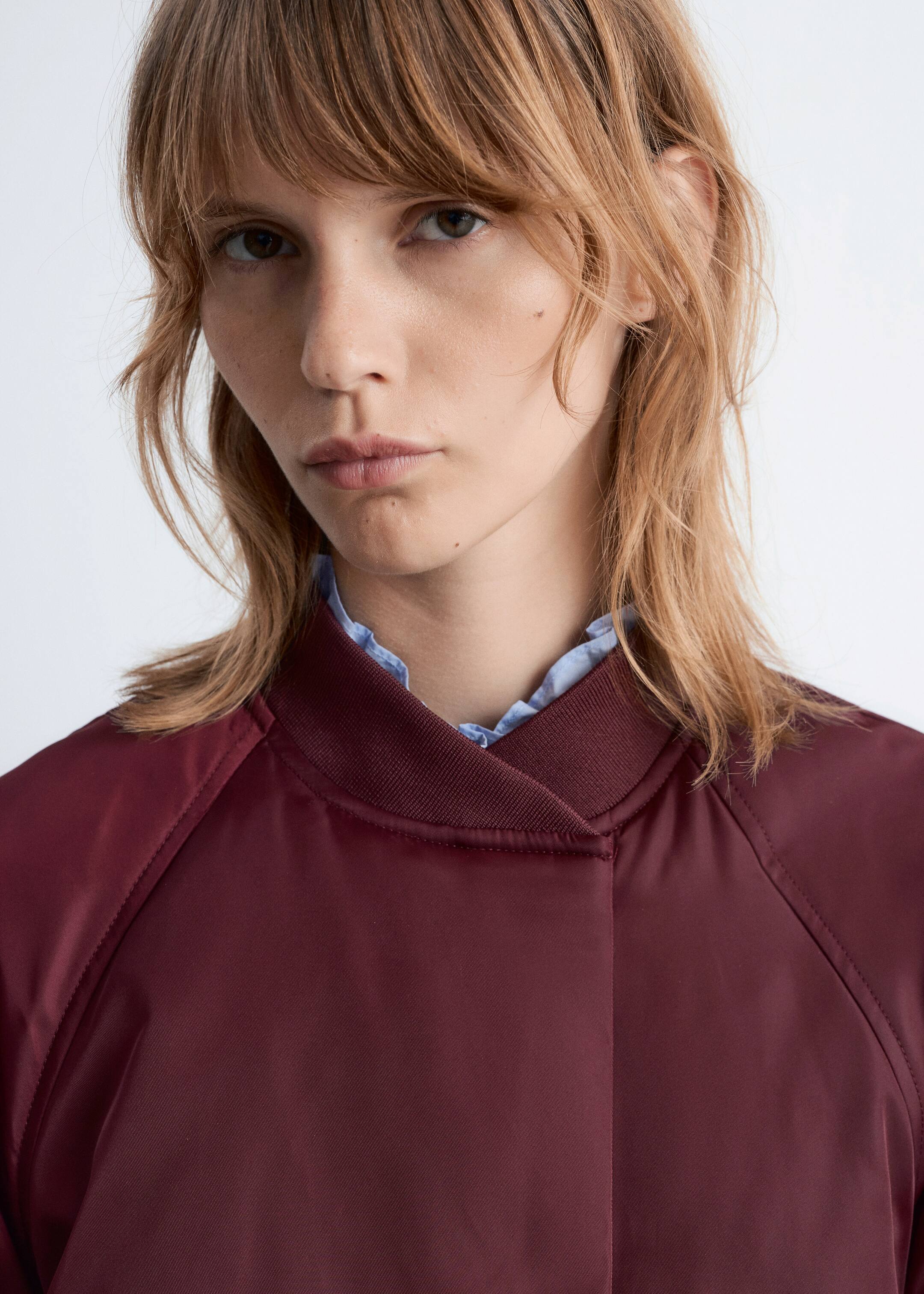 Visualizza immagine più grande: Una giacca bomber bordeaux è indossata, caratterizzata da un colletto a coste rialzato e cuciture raglan visibili. Una camicia azzurra con colletto arricciato è stratificata sotto, spuntando dal colletto della giacca.