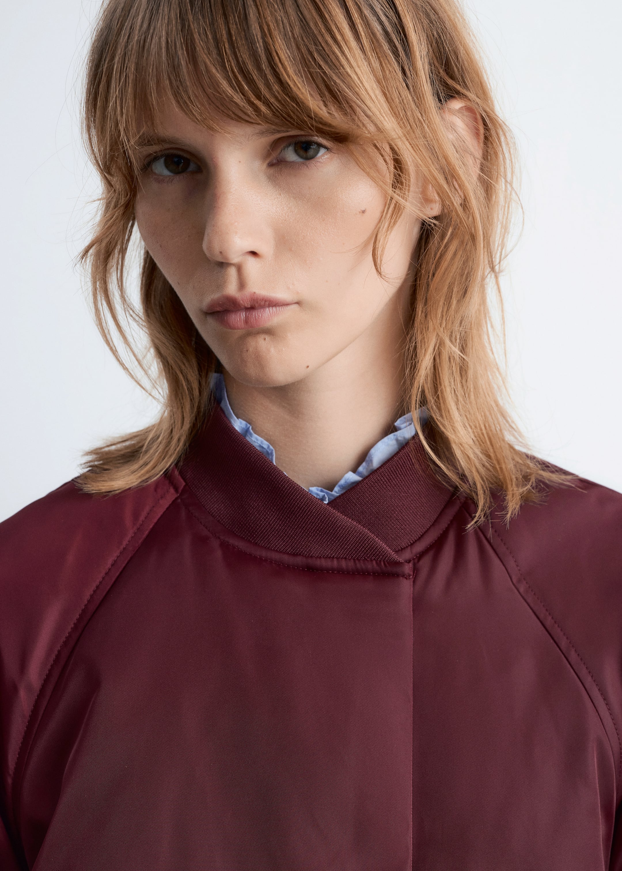 Visualizza immagine più grande: Una giacca bomber bordeaux è indossata, caratterizzata da un colletto a coste rialzato e cuciture raglan visibili. Una camicia azzurra con colletto arricciato è stratificata sotto, spuntando dal colletto della giacca.