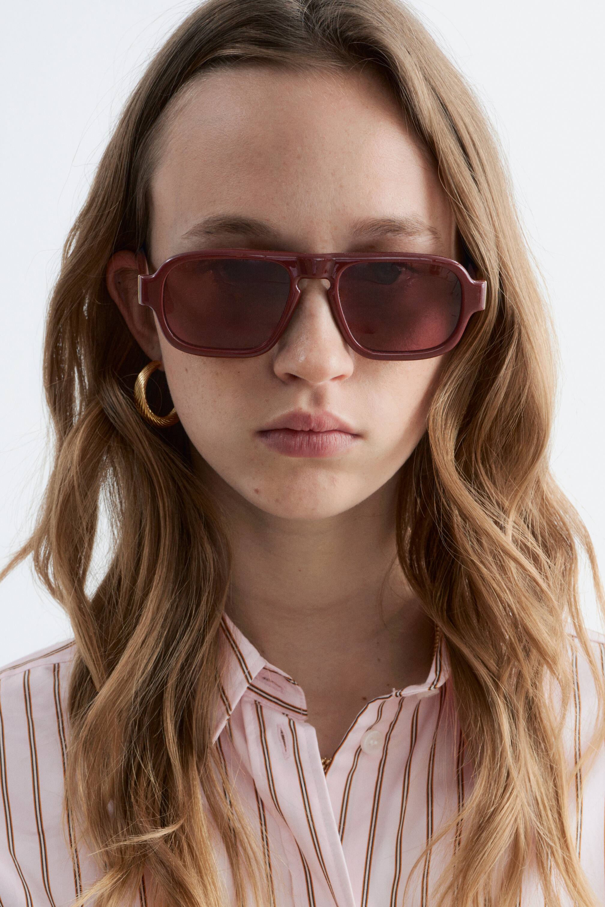 Grotere afbeelding bekijken: De zonnebril heeft een dik, bordeauxrood, rechthoekig montuur met een dubbele brug en donker roodbruin getinte glazen.