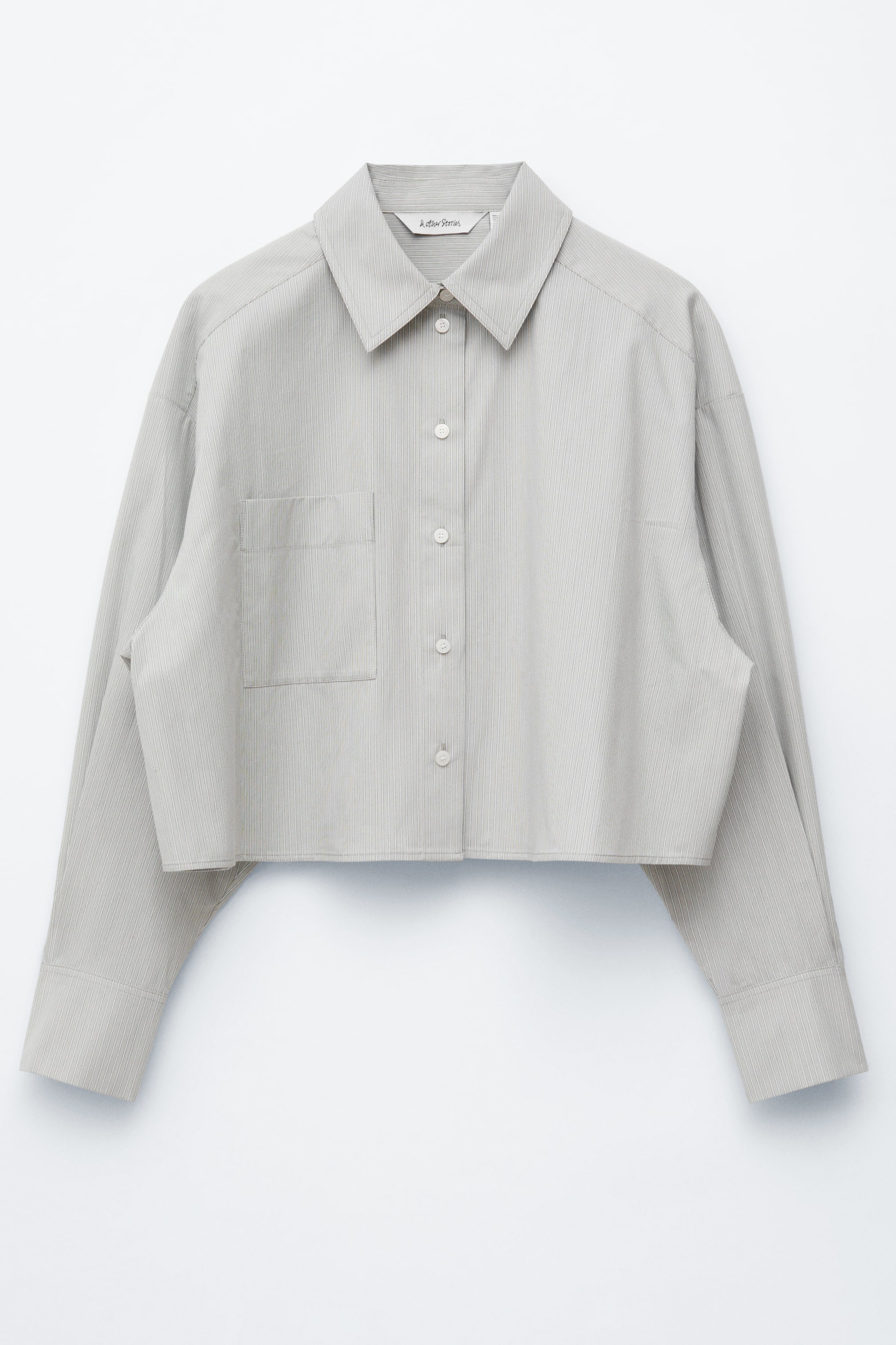 Ver imagem maior: Uma camisa cropped cinza-claro é mostrada de FRENTE, apresentando uma textura vertical fina e um silhueta oversized. Possui uma gola clássica dobrável, uma carcela abotoada, um bolso de patch à esquerda e mangas compridas com punhos abotoados.