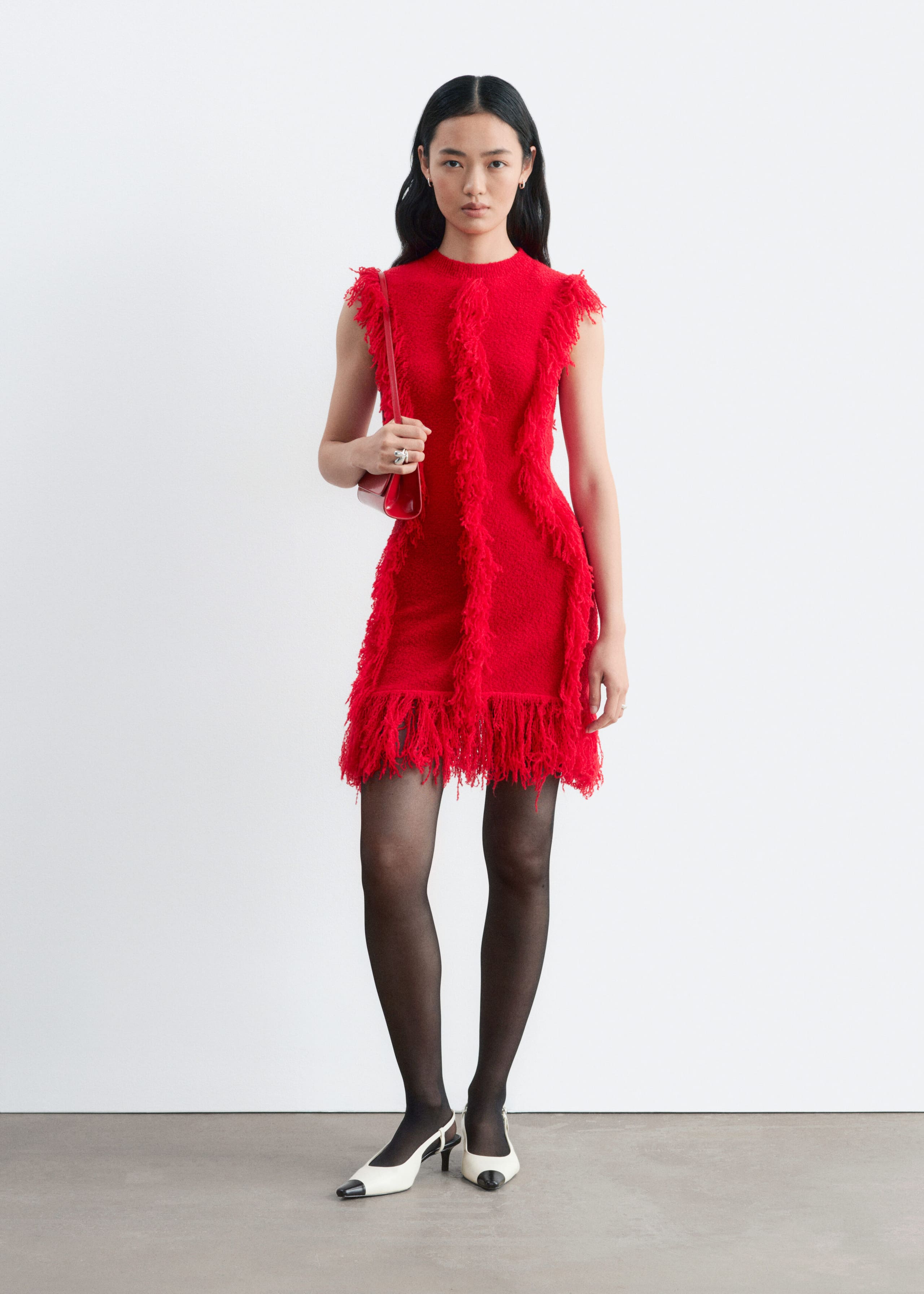 A red, sleeveless, slim-fit knit dress featuring vertical rows of fringe and a fringed hem is being worn by the model, who is also carrying a matching red shoulder bag. The outfit is styled with sheer black tights and white slingback heels with black toe caps.