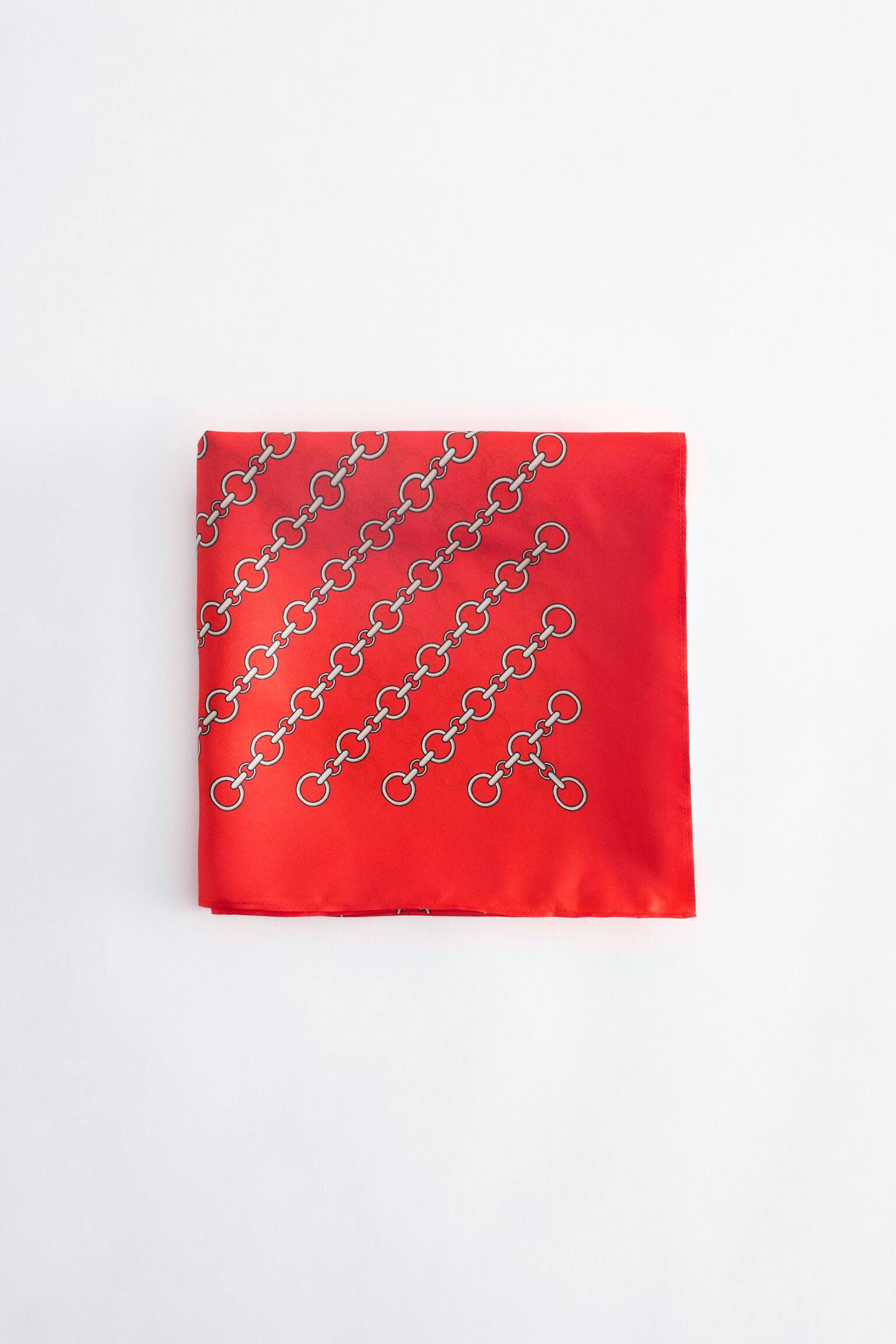 Una sciarpa di seta rossa piegata a quadrato mostra un motivo diagonale di maglie di catena in tonalitÃ  argento sulla sua superficie liscia. Il prodotto Ã¨ presentato frontalmente, mettendo in risalto la sua finitura satinata e i bordi puliti.
