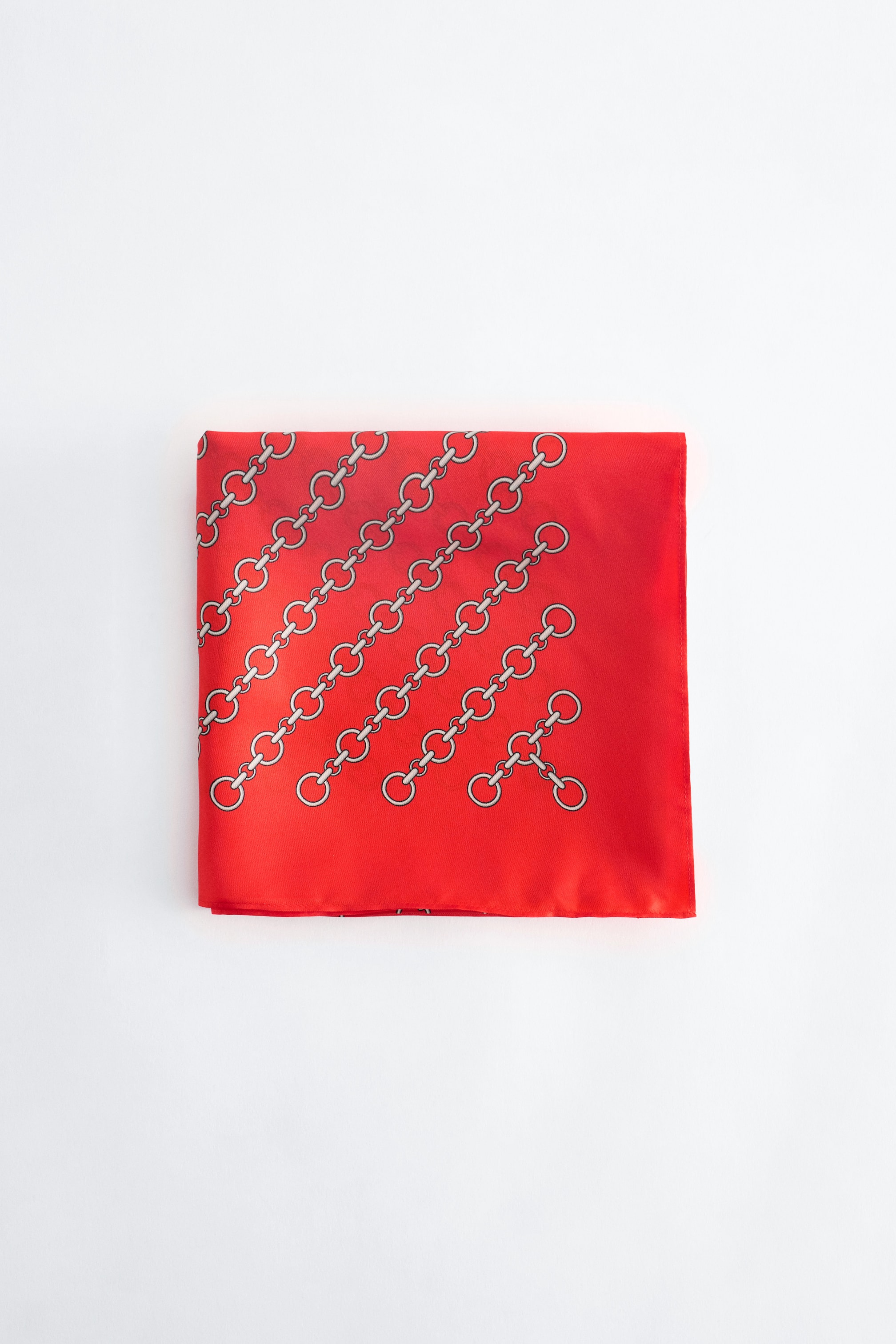 Visualizza immagine più grande: Una sciarpa di seta rossa piegata a quadrato mostra un motivo diagonale di maglie di catena in tonalità argento sulla sua superficie liscia. Il prodotto è presentato frontalmente, mettendo in risalto la sua finitura satinata e i bordi puliti.