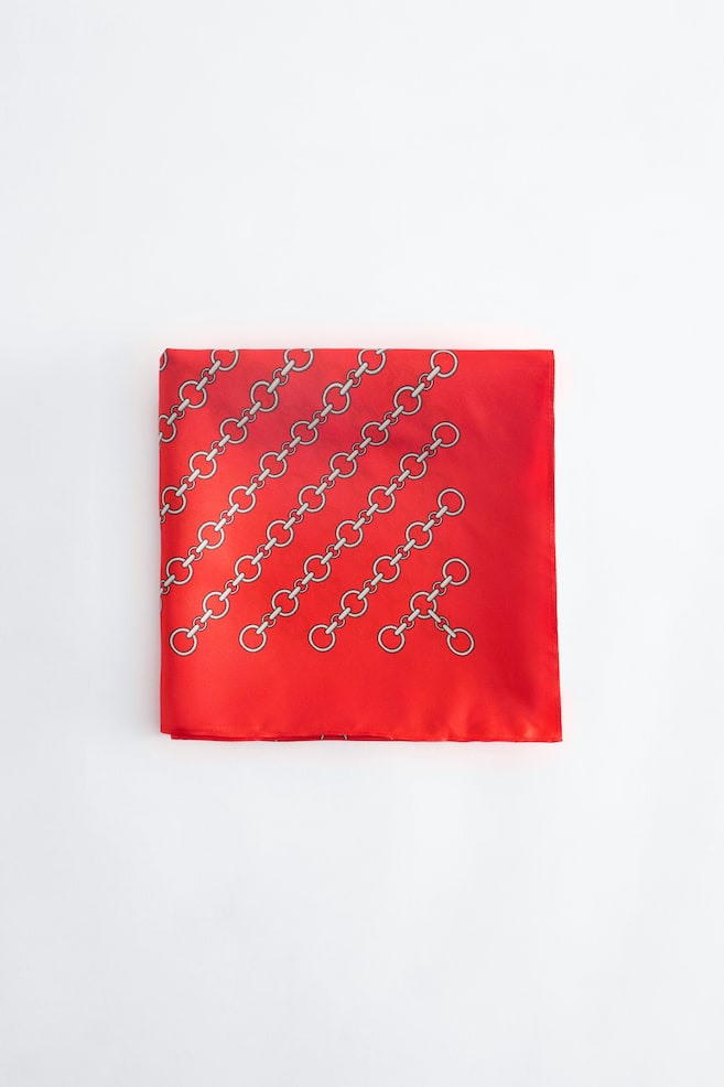 Une écharpe en soie rouge, pliée en carré, présente un motif diagonal de maillons de chaîne aux reflets argentés sur sa surface lisse. Le produit est présenté de face, mettant en valeur son fini satiné et ses bords nets. - 1