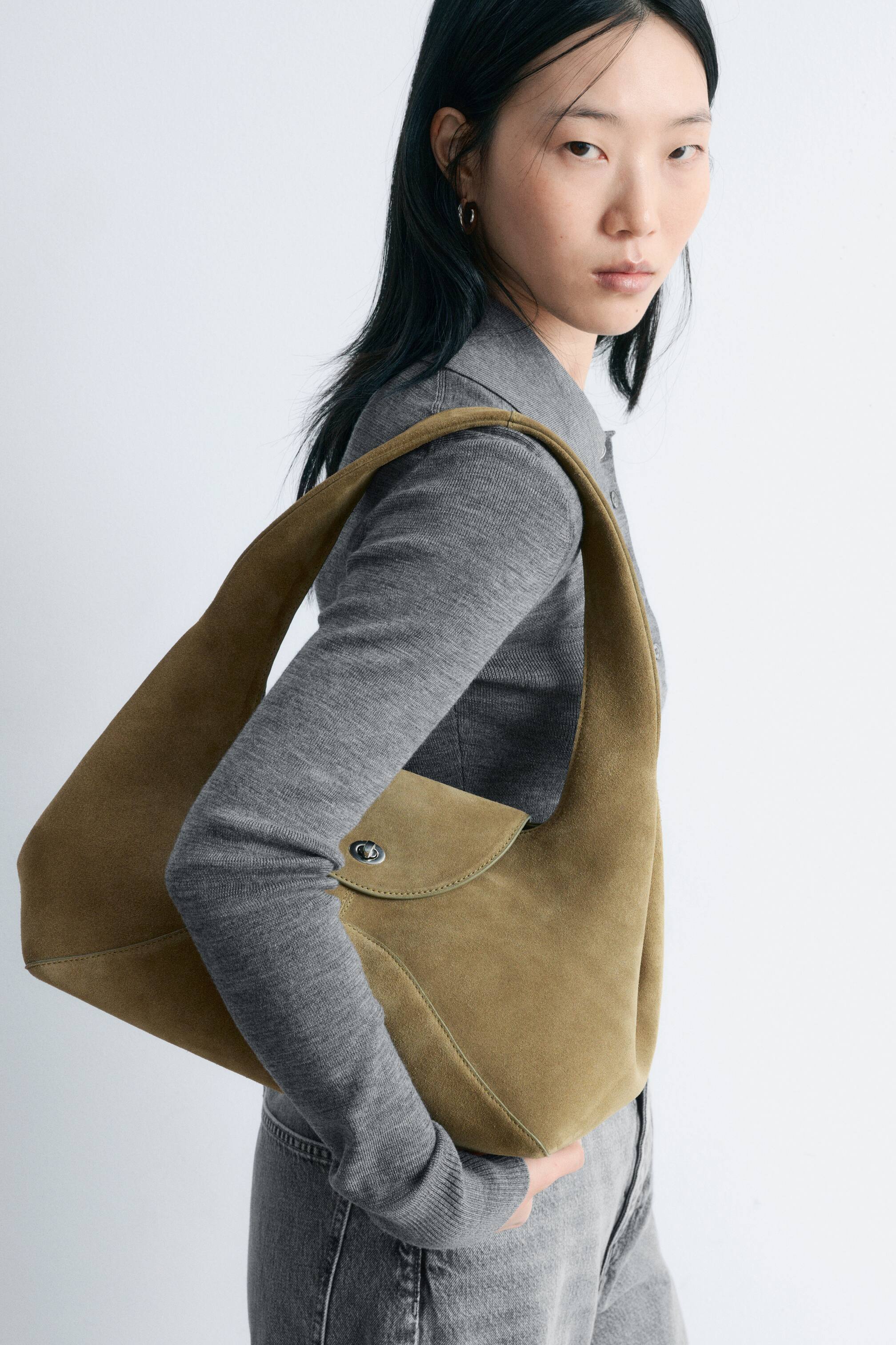 Vezi imaginea mai mare: O geantă de umăr din piele întoarsă, de culoare maro-oliv, cu o siluetă curbată și sculpturală, o curea lată și înclinată, o cusătură decorativă în linie dreaptă și o clapetă frontală cu închidere prin răsucire, este purtată pe umărul modelului.