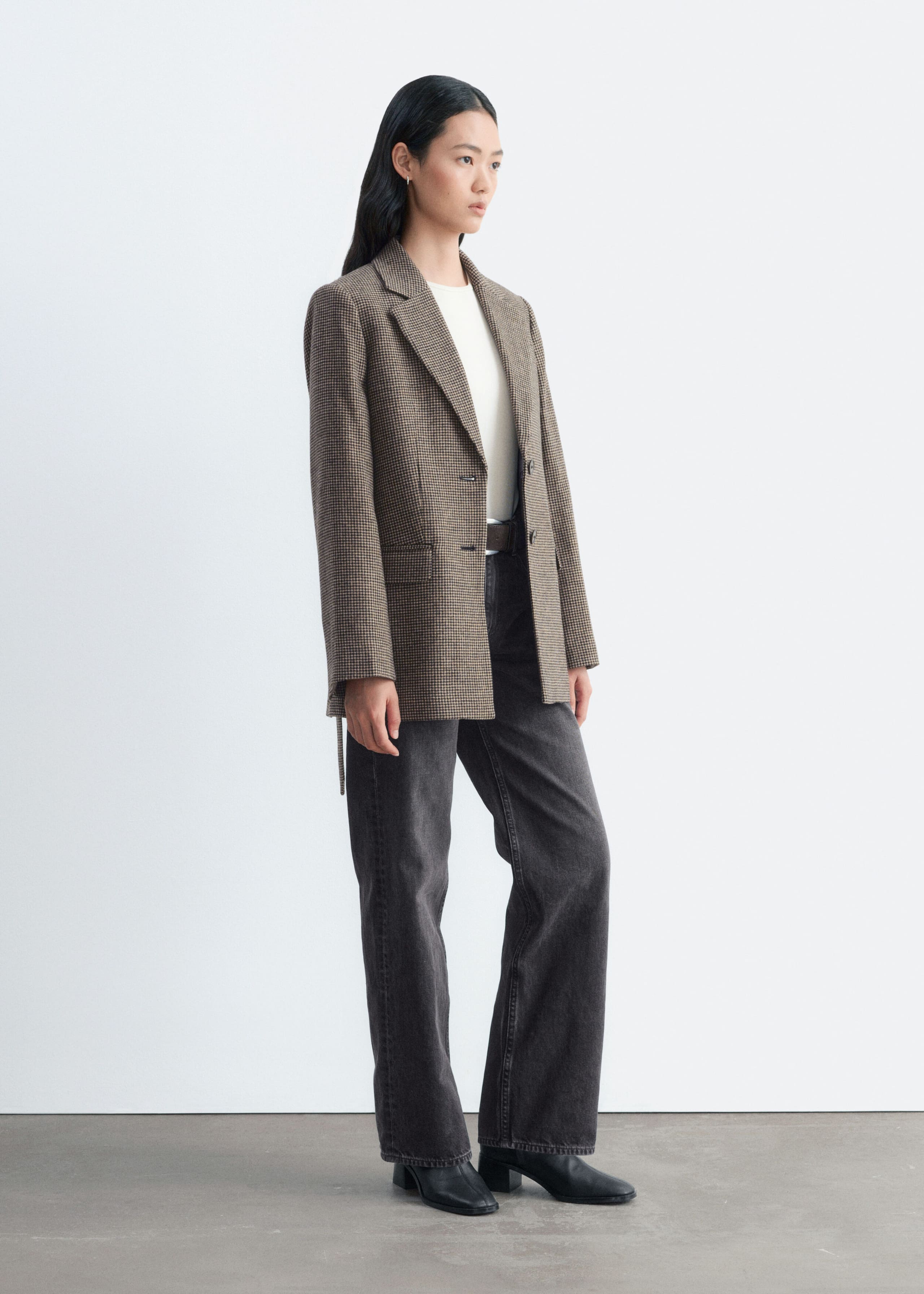 A brown houndstooth wool-blend blazer is worn open by the model, featuring notched lapels, flap pockets, and a loose tie belt hanging at the side. It is styled over a white T-shirt and paired with wide-leg dark grey jeans and black boots.
