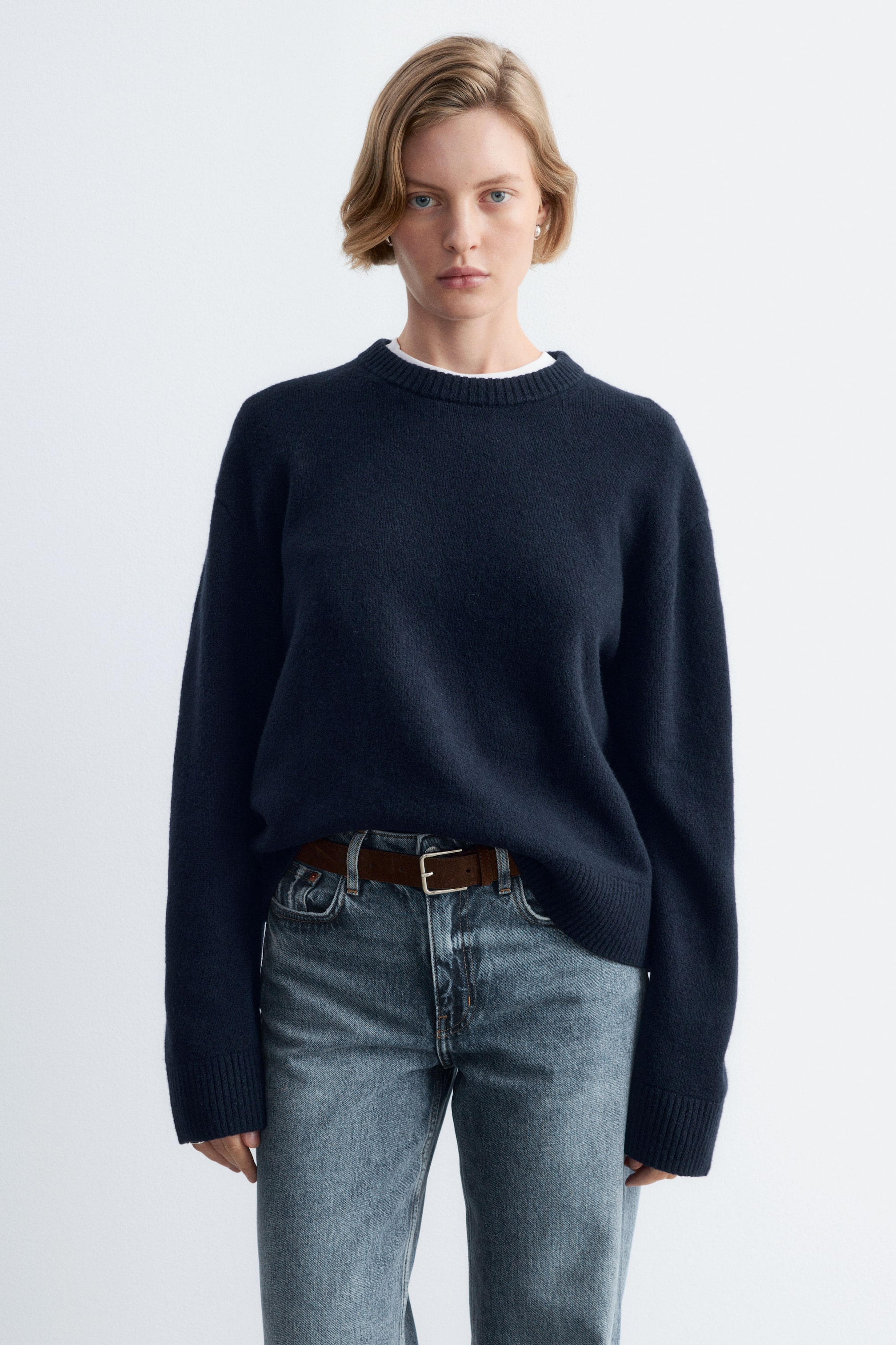 Visualizza immagine più grande: La modella indossa un maglione lavorato a maglia blu navy dal taglio boxy, con girocollo a coste e maniche lunghe con polsini a coste. Il maglione è indossato sopra una T-shirt bianca ed è infilato morbidamente sul davanti nella vita dentro un paio di jeans blu, abbinati a una cintura marrone.