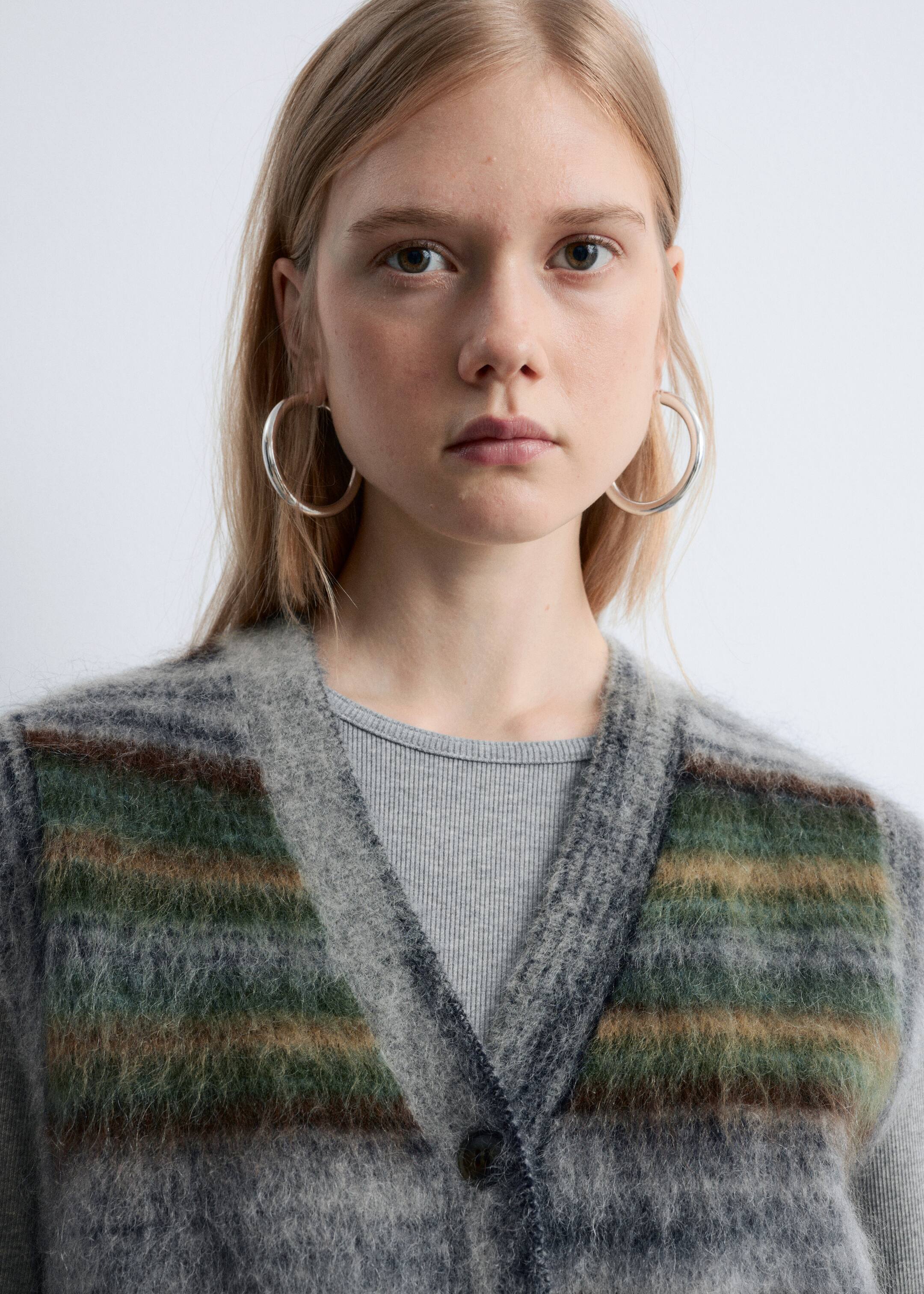 Visualizza immagine più grande: Un cardigan lavorato a maglia in grigio viene presentato frontalmente, caratterizzato da uno scollo a V e strisce orizzontali in verde tenue e marrone. È indossato sopra un top a coste grigio chiaro, con un bottone scuro visibile all'abbottonatura anteriore.