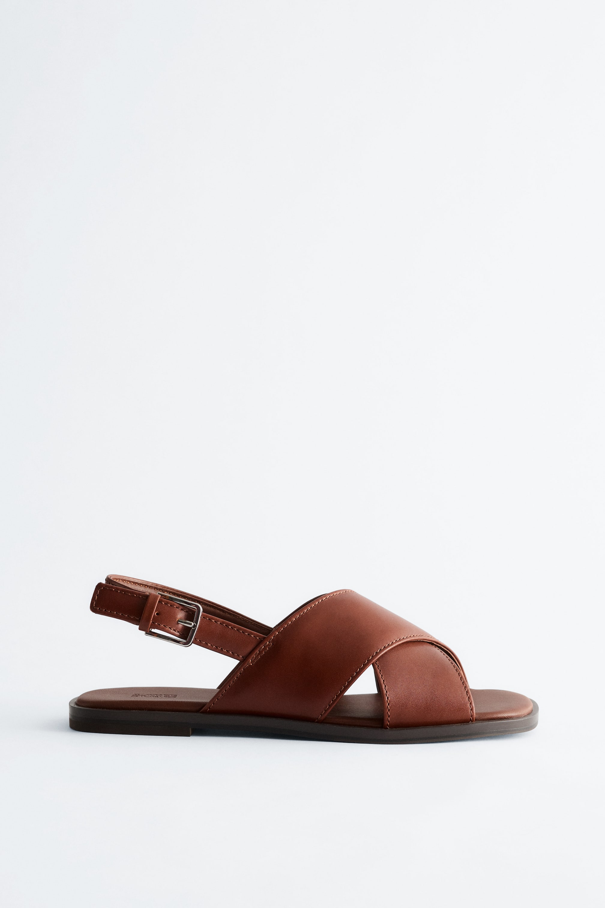 Ver imagem maior: Uma sandália de couro castanha está virada para a esquerda, apresentando uma tira larga em cruz sobre o peito do pé, uma tira no tornozelo com fivela de tom prateado e uma sola plana de castanho escuro.