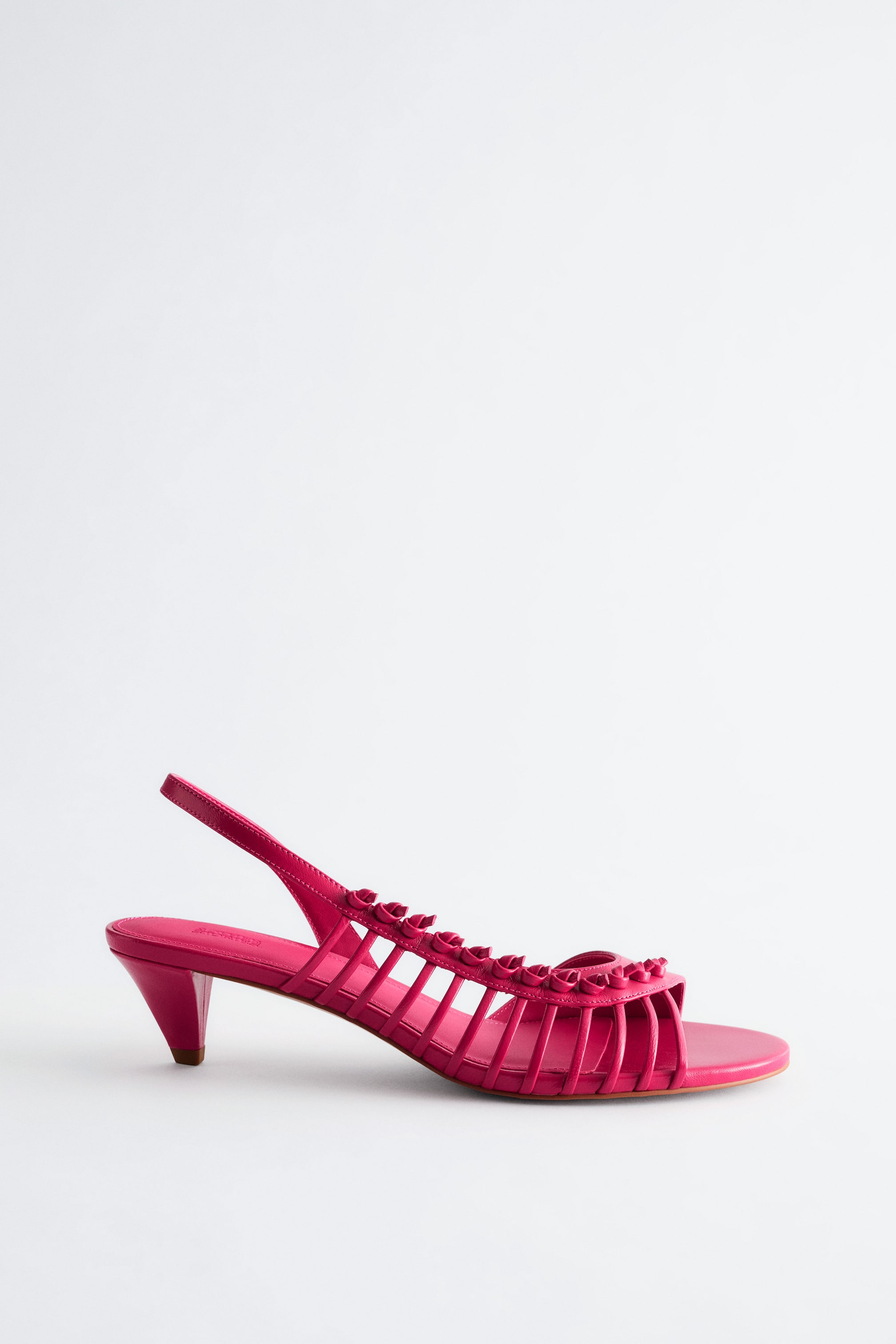 Agrandir l'image: Une seule sandale slingback en cuir rose fuchsia est orientée vers la gauche. La sandale présente un talon aiguille, une bride arrière et un design de tige en cage avec des brides décoratives à volants.