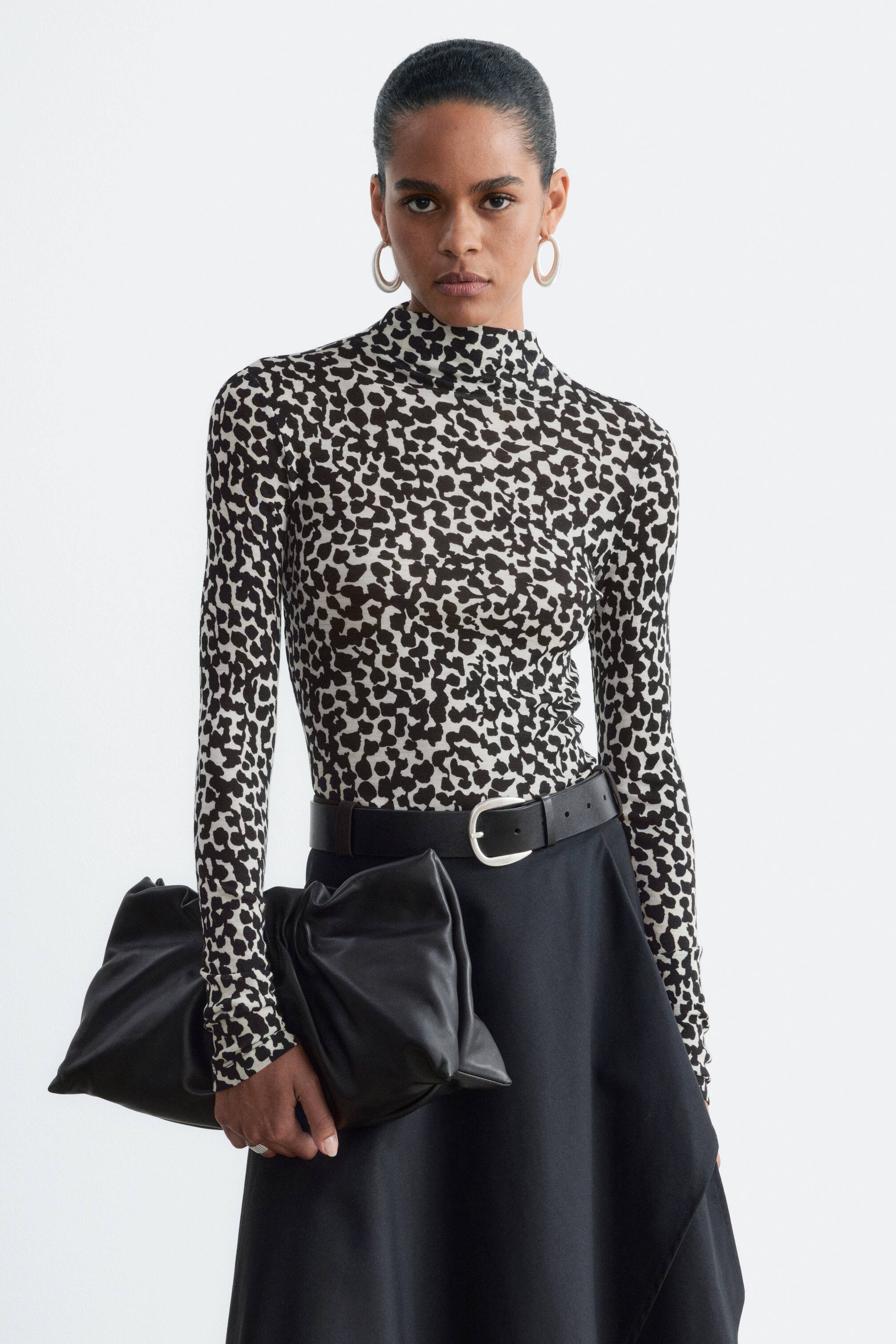 View larger image: A wool mock-neck top with a black and white abstract animal print is worn by the model, tucked into a black skirt with a belted waist. The long-sleeved, fitted top is styled with a large black leather clutch held in the model's hand.