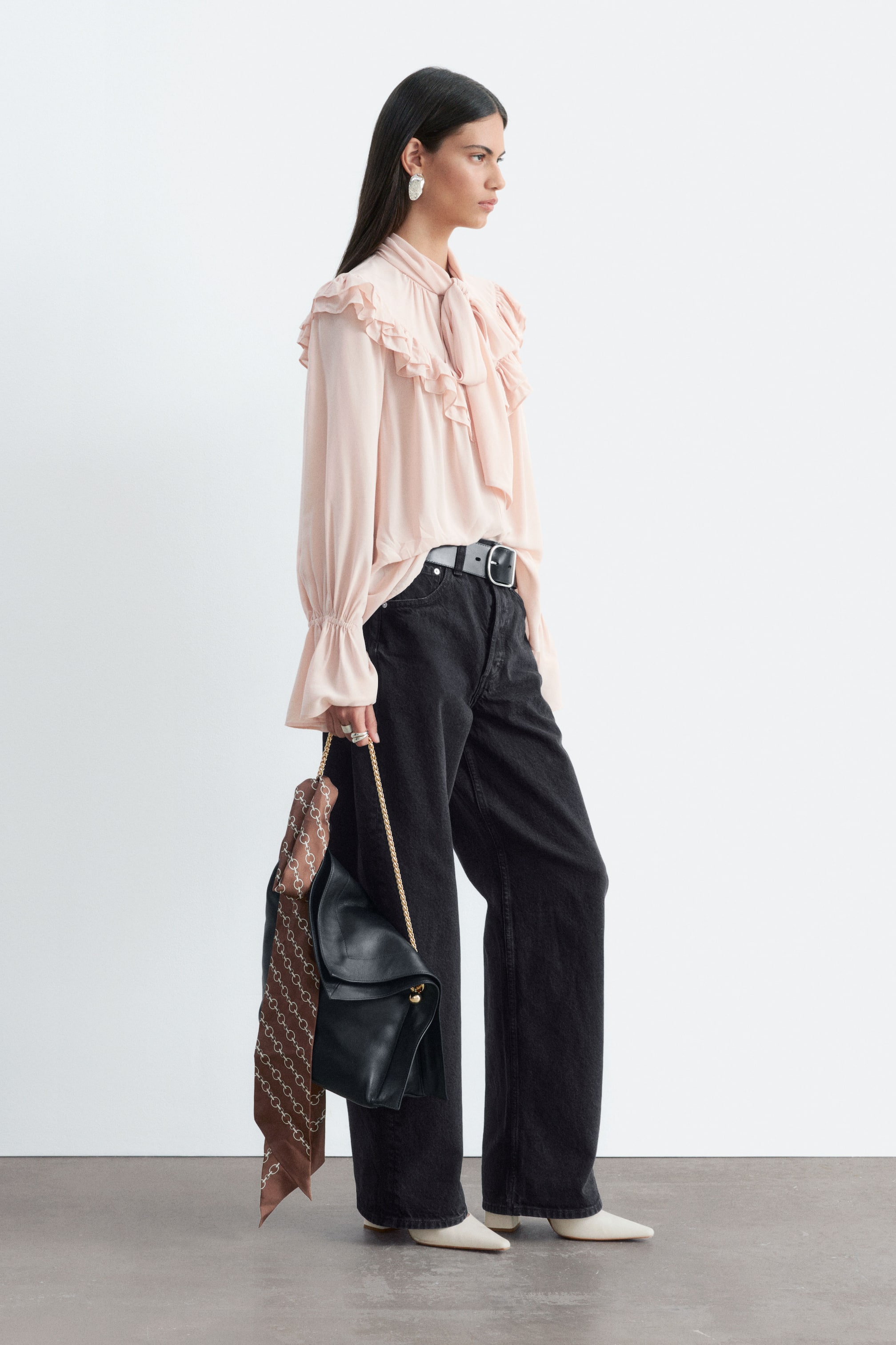 Agrandir l'image: Le chemisier rose pâle à détail foulard et volants est porté par le mannequin. Il présente un col foulard noué, des détails à volants sur les épaules et des manches longues à poignets évasés. Le chemisier est porté rentré dans un jean noir à jambe large, maintenu par une ceinture en cuir noir.
