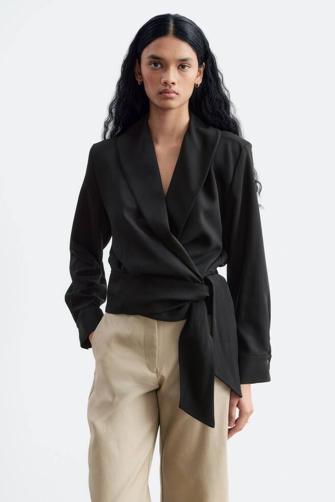 En svart omslagsbluse med myk sjalkrage, lange ermer og knyting i front ved livet bÃ¦res av modellen. Blusen er stylet med et par beige bukser. - 1