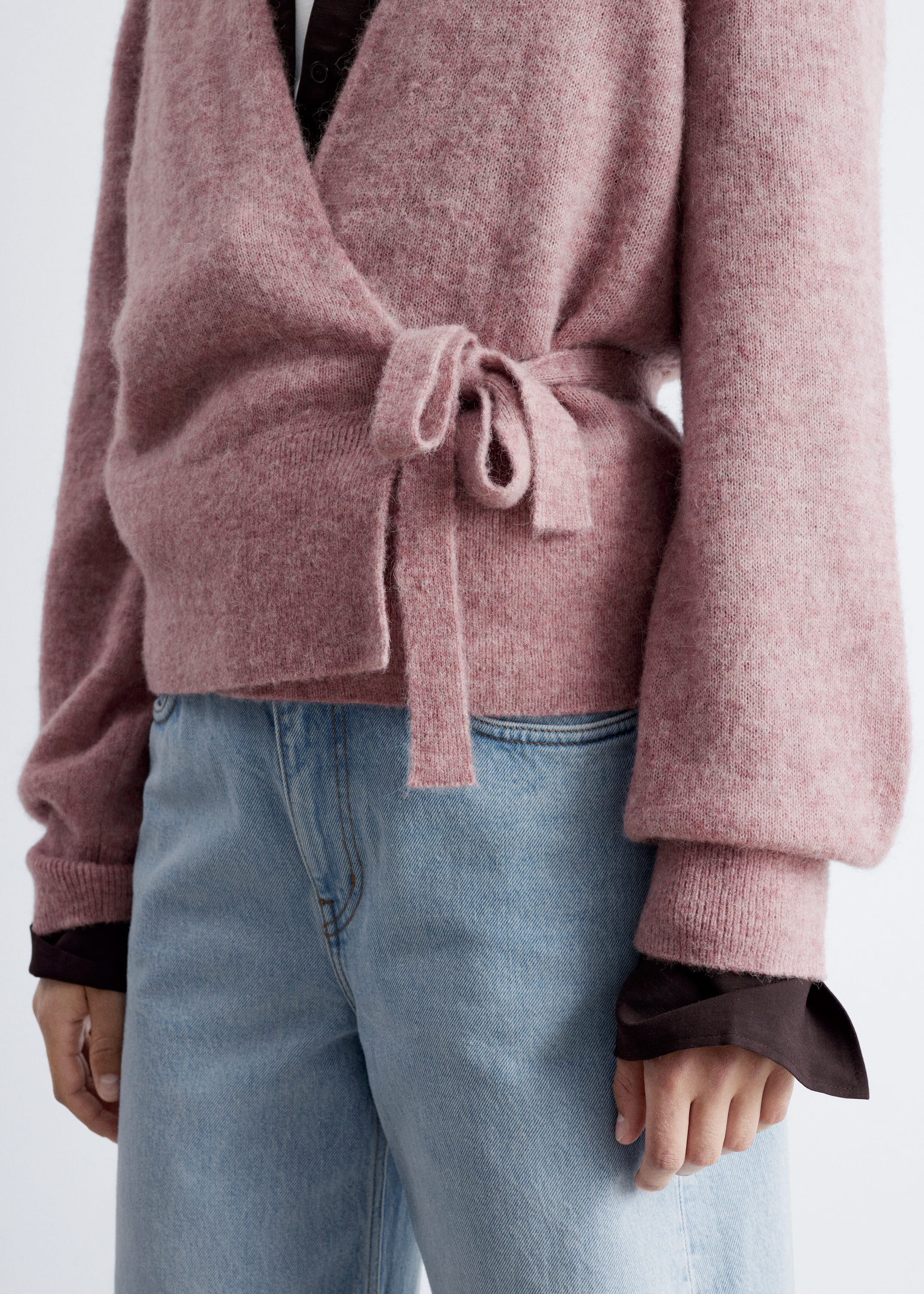 Ver imagem maior: Um casaco de malha rosa com detalhe de amarração na cintura, mangas compridas bufantes e punhos e bainha canelados está a ser usado pela modelo. A modelo está a usar o casaco sobre um top castanho escuro e está a usar jeans azul claro.