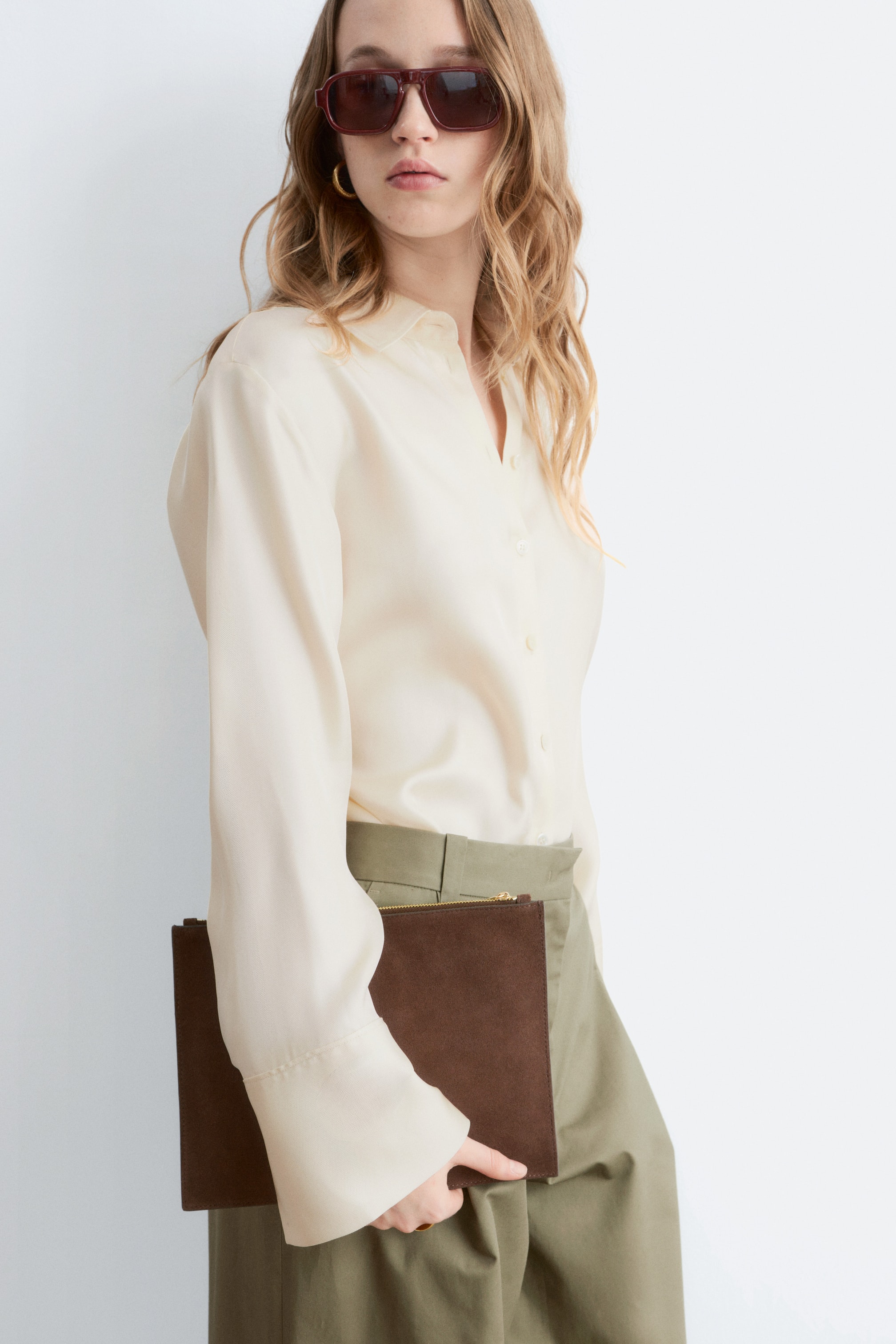 Ingrandisci l'immagine: Una pochette rettangolare in camoscio marrone con una cerniera dorata viene tenuta accanto al fianco della modella.