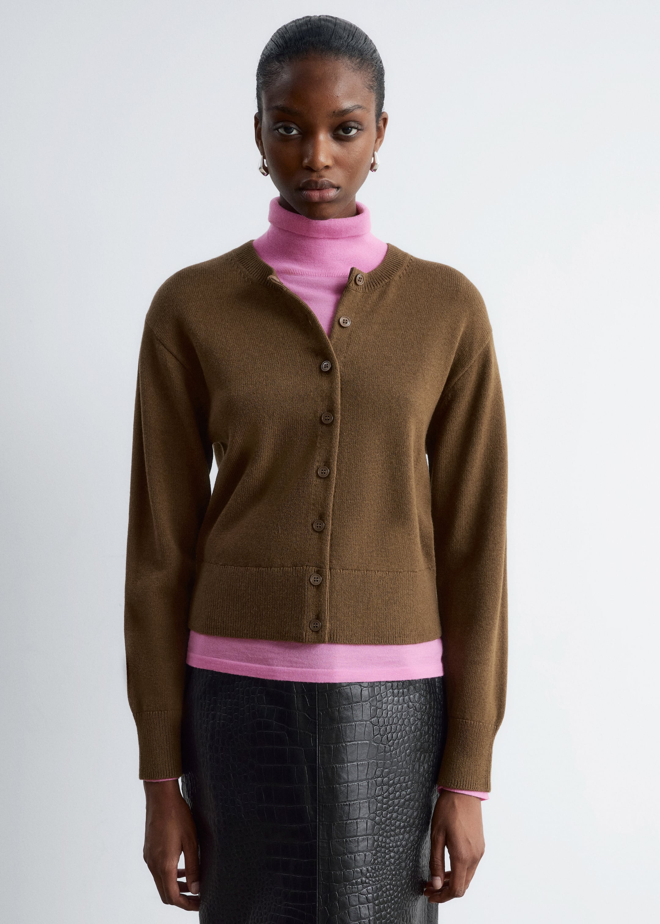 Grösseres Bild ansehen: Eine braune Strickjacke mit Rundhalsausschnitt wird getragen, vorne zugeknöpft, über einem rosa Rollkragenpullover und einem schwarzen Rock. Sie verfügt über sechs runde braune Knöpfe, lange Ärmel und gerippte Bündchen und Saum und bietet eine lockere Passform.