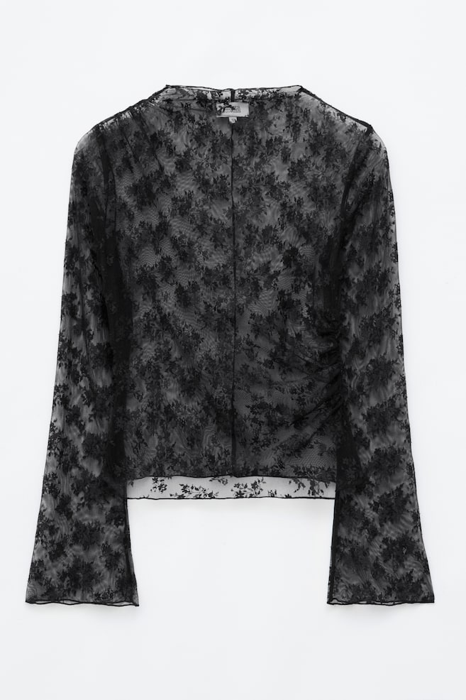 Un top de encaje negro presentado de frente, que muestra un estampado floral semitransparente, cuello alto redondo y mangas largas acampanadas con remates baby lock. El top presenta fruncido asimétrico en un hombro y en la costura lateral opuesta, creando una silueta drapeada. - 1