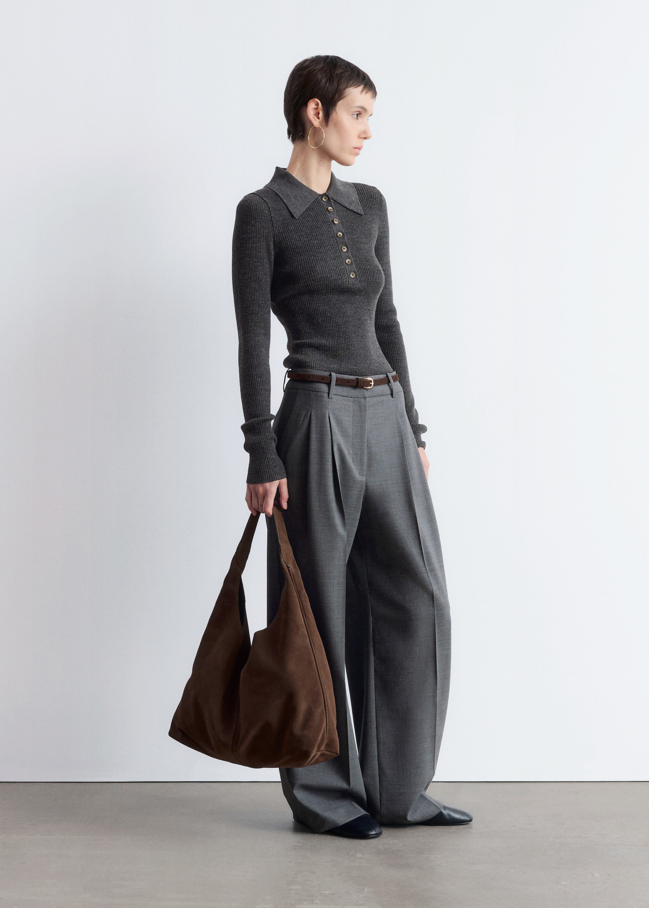 View larger image: A brown suede tote bag with a matte finish is being carried by the model. The bag has a soft, unstructured shape and is held by its wide, integrated strap.