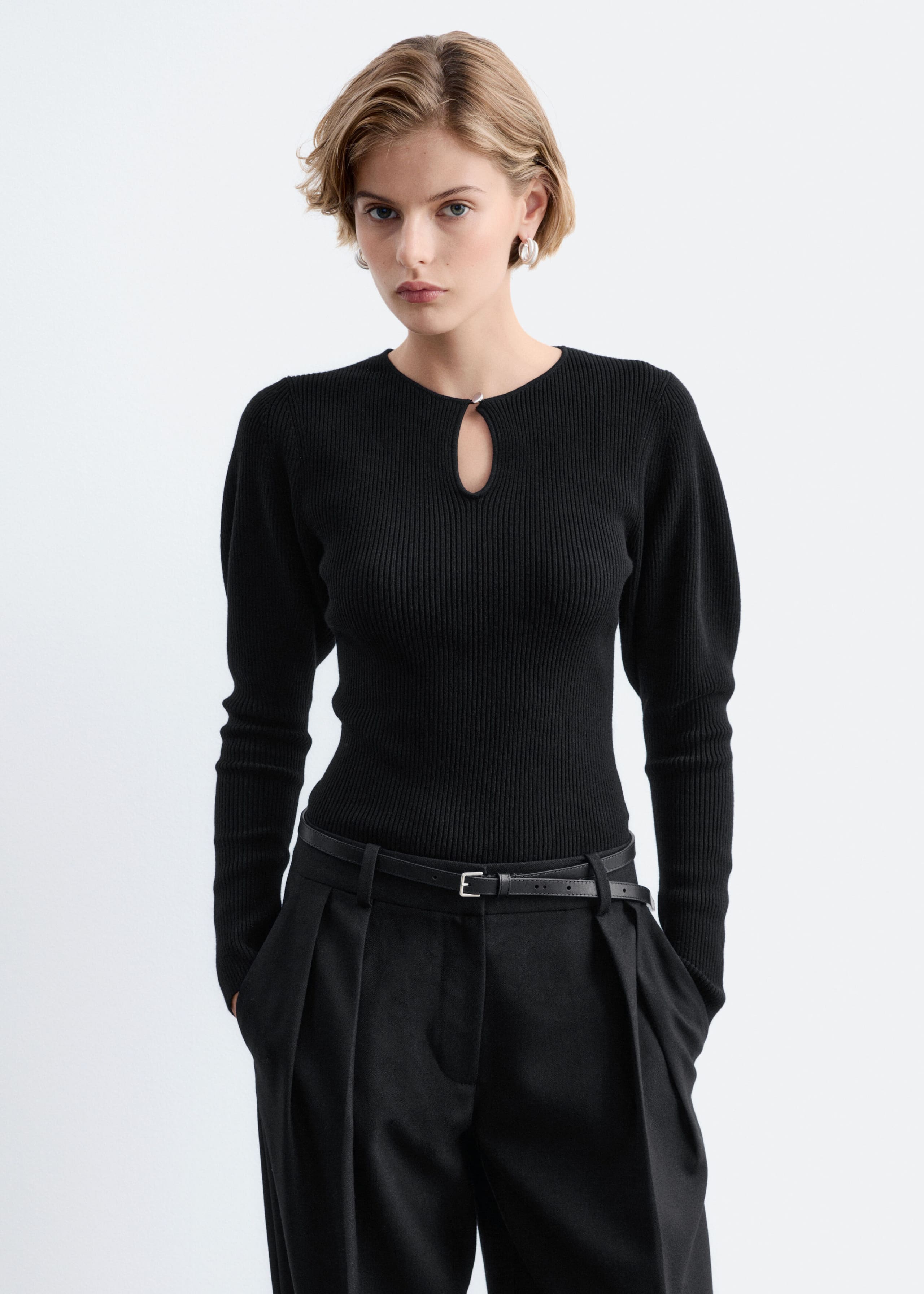 A black rib-knit top with a front keyhole opening and softly puffed shoulders is being worn by the model. The fitted, long-sleeved top is tucked into black pleated trousers with a slim black belt.