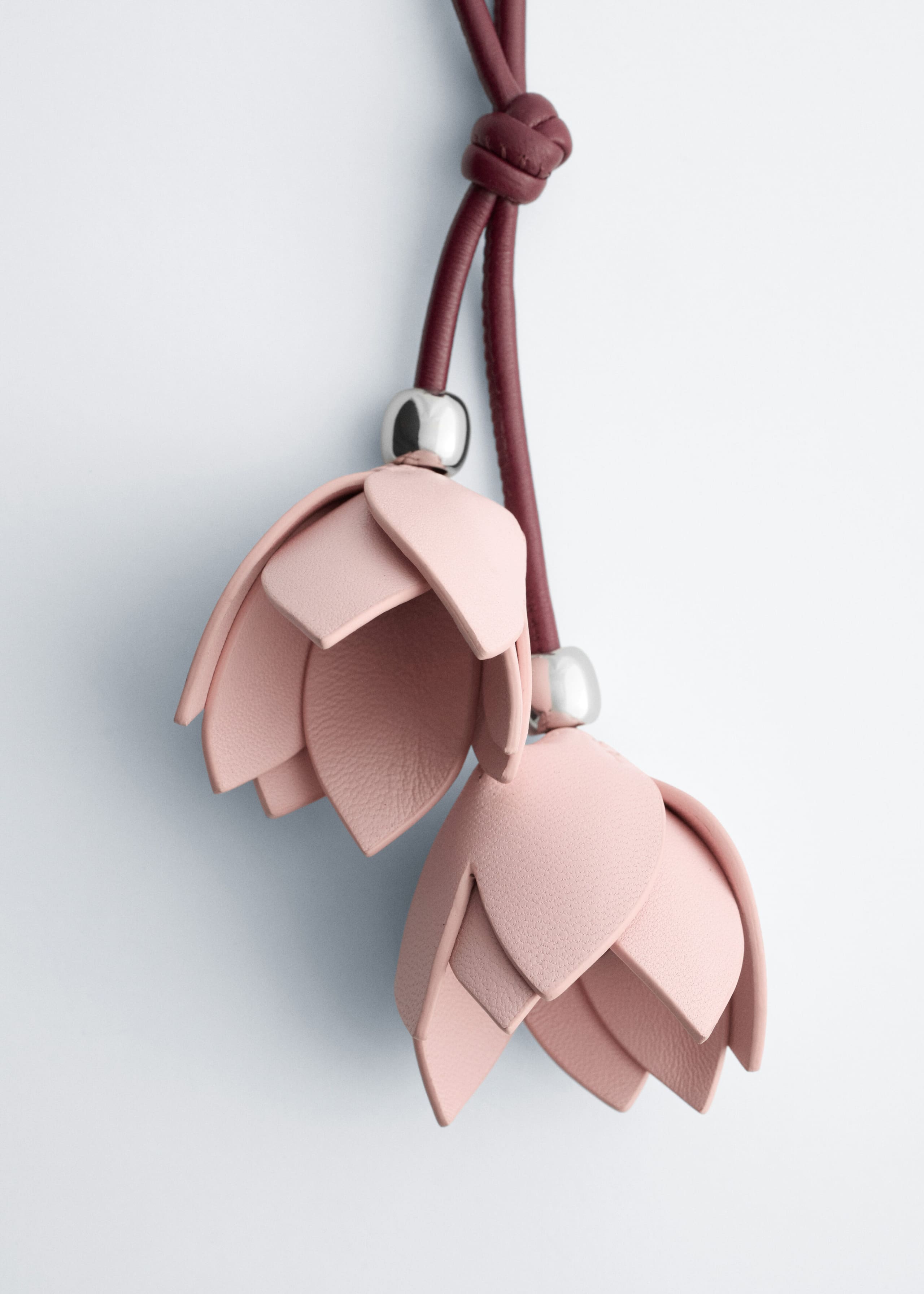 The flower bag charm features two pale pink leather flower pendants shaped like tulips, hanging at staggered lengths from a knotted burgundy leather cord. Each flower features layered, textured petals and is topped with a polished silver-tone metallic bead.