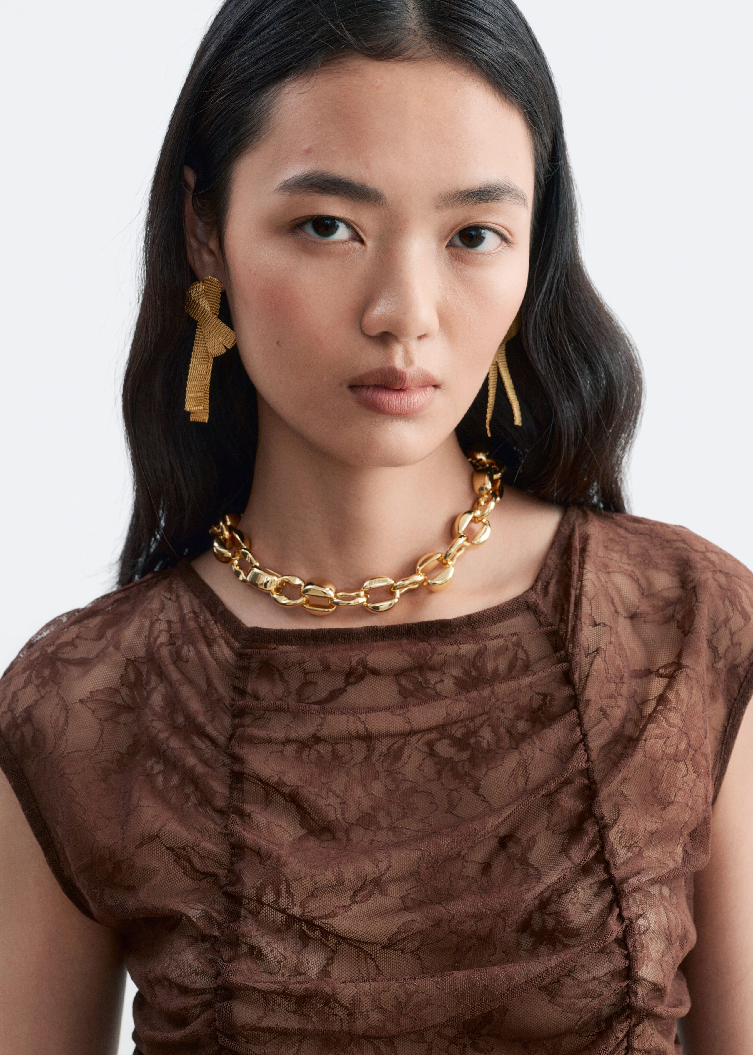 A brown lace top featuring a floral pattern, vertical ruching, and a boat neckline is worn by the model. The top is styled with a chunky gold chain necklace and gold bow-shaped earrings.